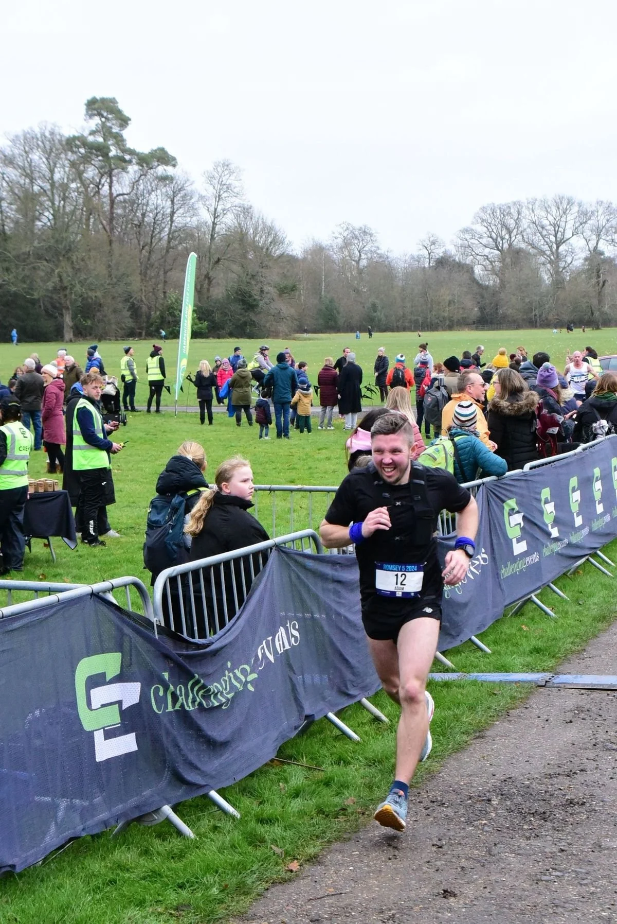 Adam Abel crossing the finish line during a race, smiling and wearing a black athletic outfit, with spectators and race officials behind a barrier on a grassy field in a park.
