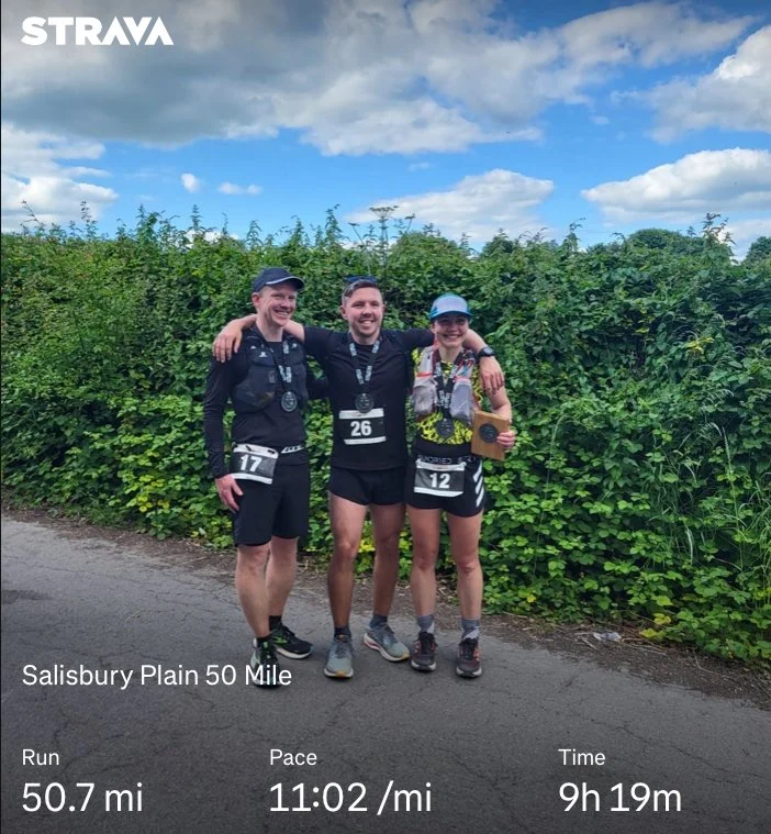 Adam Abel standing together with arms around each Ruth Barker and Patrick after completing the Salisbury Plain 50 Mile race, smiling at the camera, wearing race bibs and medals, outdoors with green bushes and partly cloudy sky in the background.