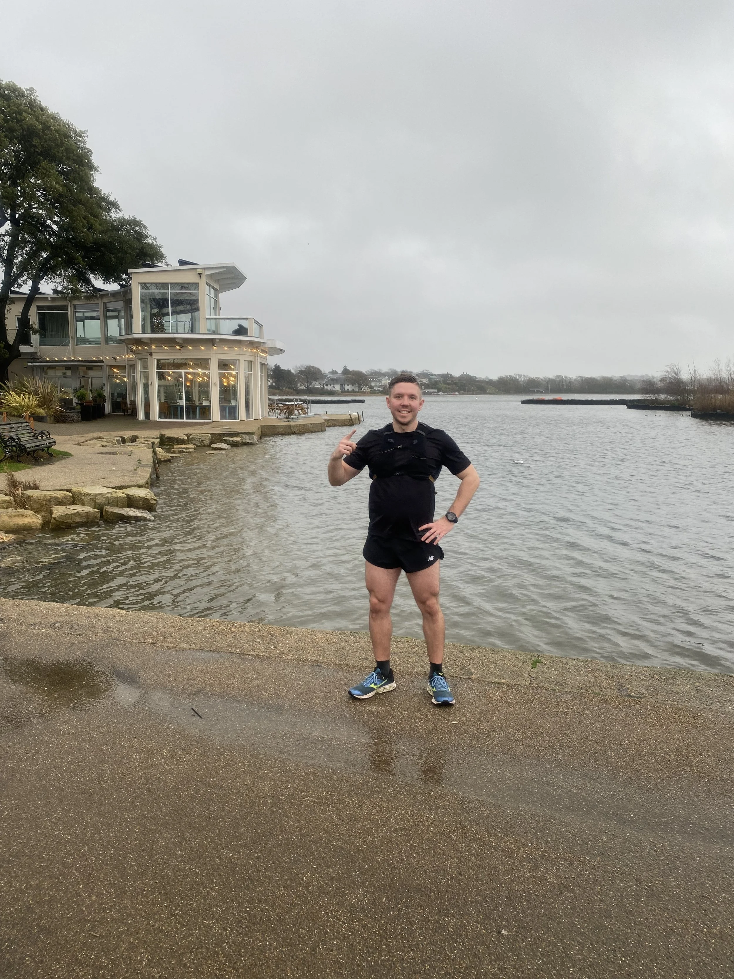Adam Abel in running shoes and athletic clothing standing by a body of water, pointing at himself, with a modern house and trees in the background on a cloudy day at Poole Parkrun having ran it in 19:29.