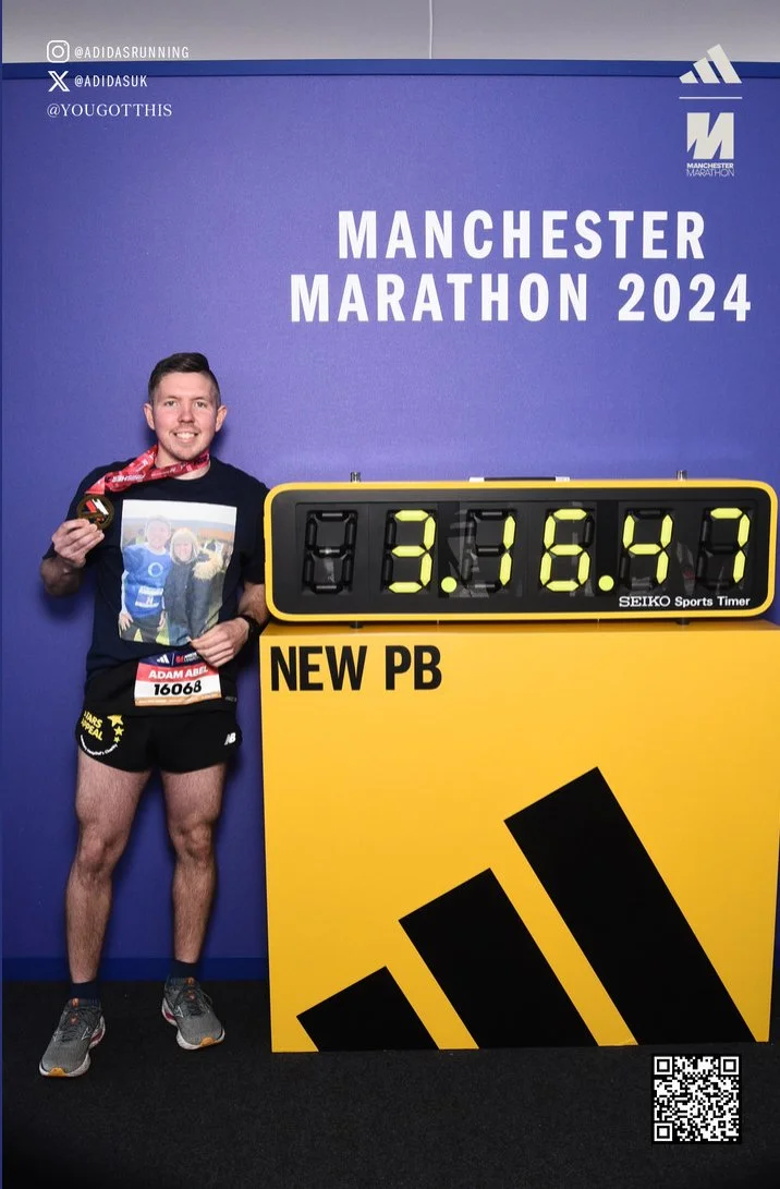 Adam Abel, marathon runner in black shirt and shorts holding a photo and a medal, standing next to a digital timer displaying 3:16:43 at the Manchester Marathon 2024 event.