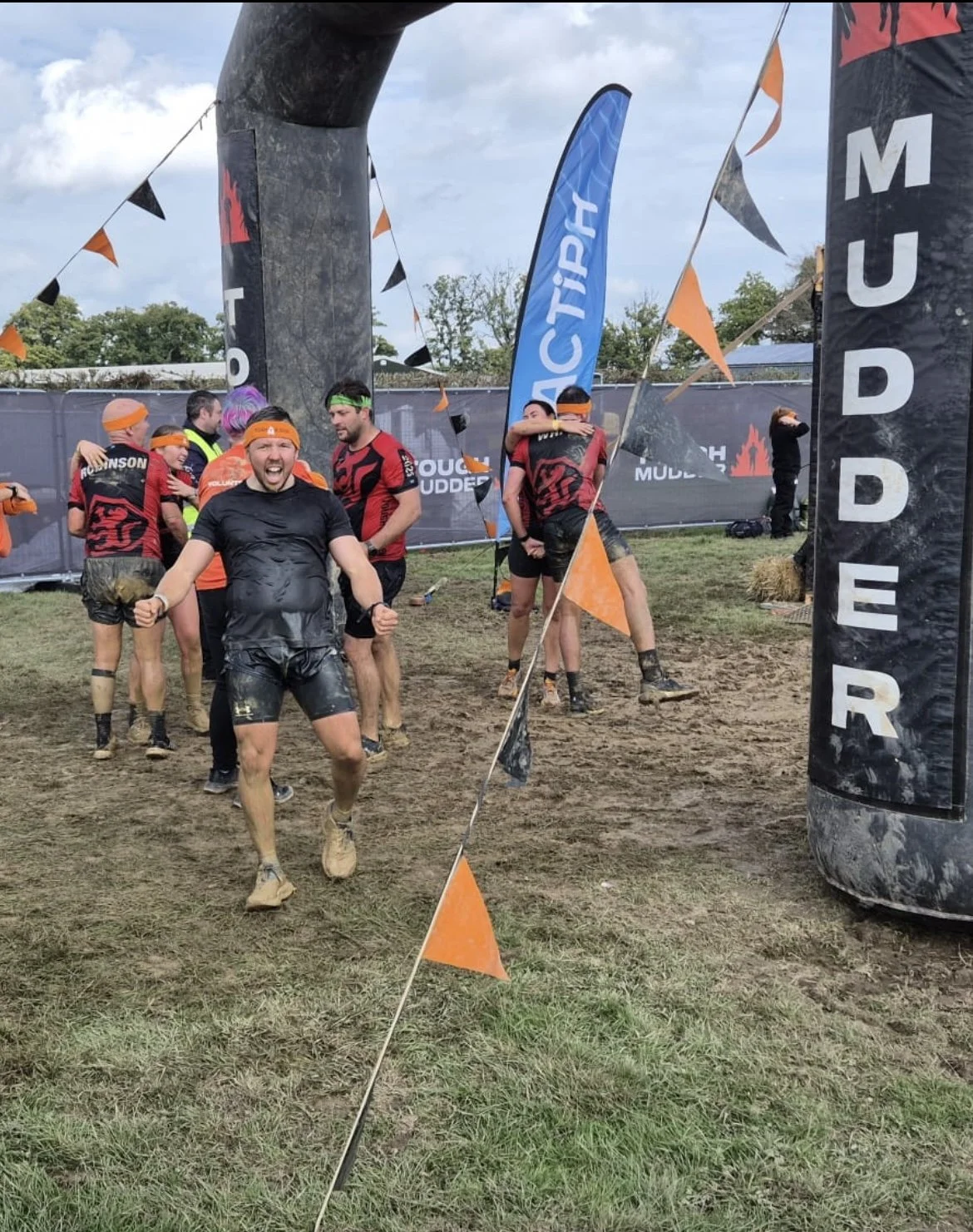 Adam Abel celebrating after completin tough mudder 15k, muddy and exhausted, with flags and banners around the finish line.