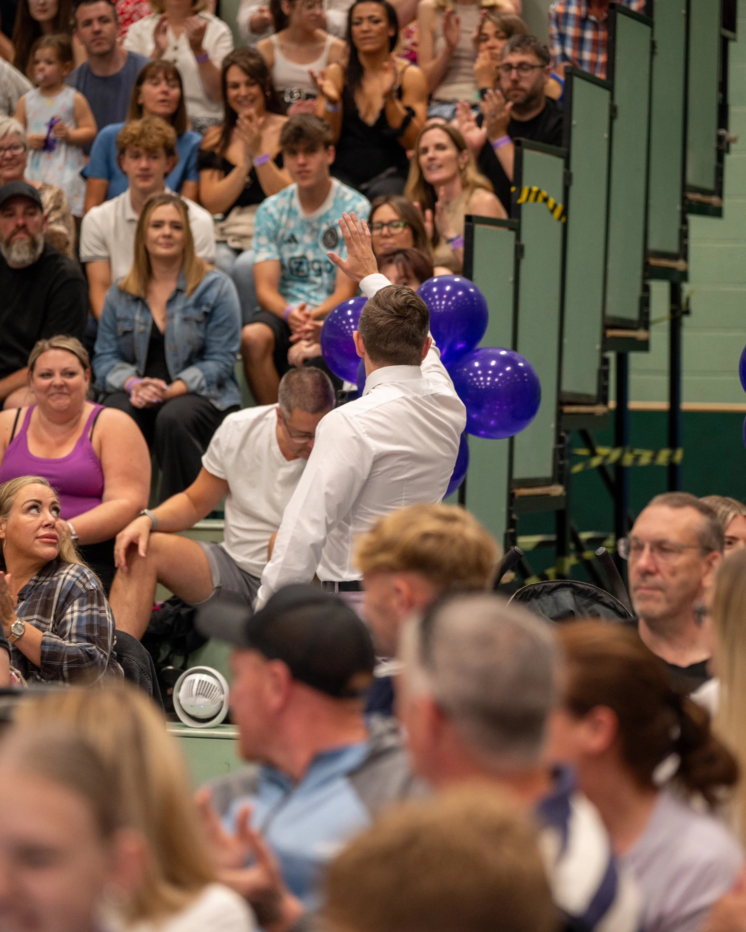 Adam Abel raising his hand to wave or address a crowd. The crowd, seated in a auditorium, watching him with interest and smiling. Blue balloons are visible, and some people are clapping.