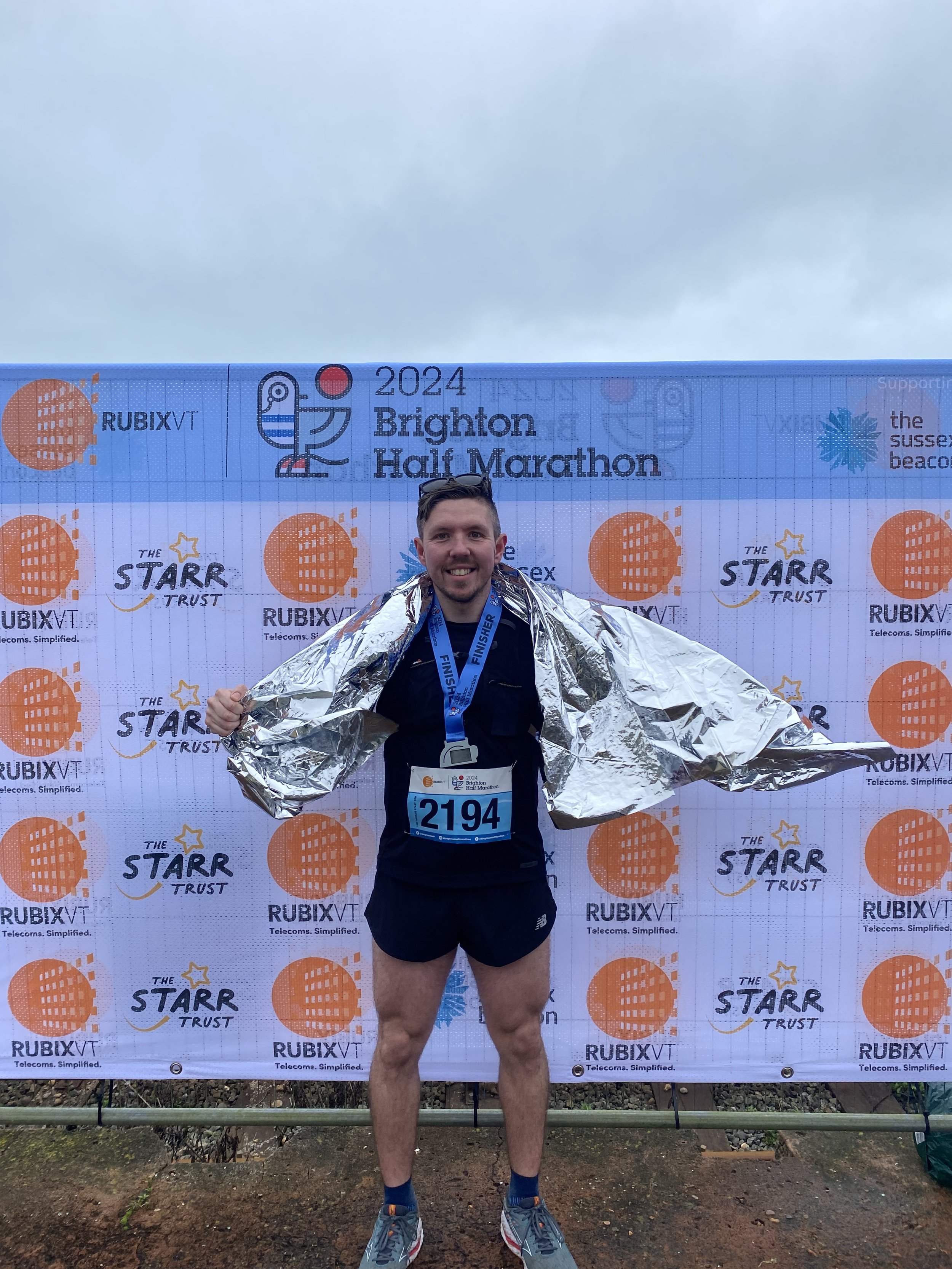 Adam Abel, wearing a finisher's medal and a metallic emergency blanket around his shoulders, standing in front of a banner at the 2024 Brighton Half Marathon after running it in 1:29:35