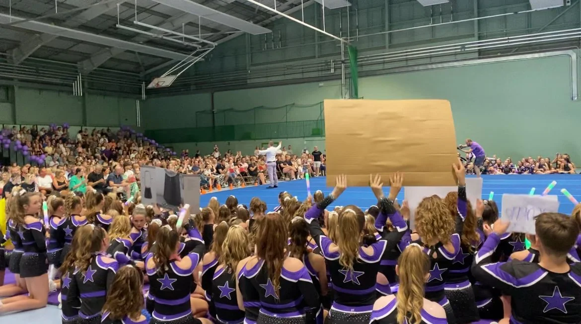 Cheerleaders sitting on the floor in a gymnasium during a cheerleading showcase or event, with audience members in the background, and Adam Abel standing on stage