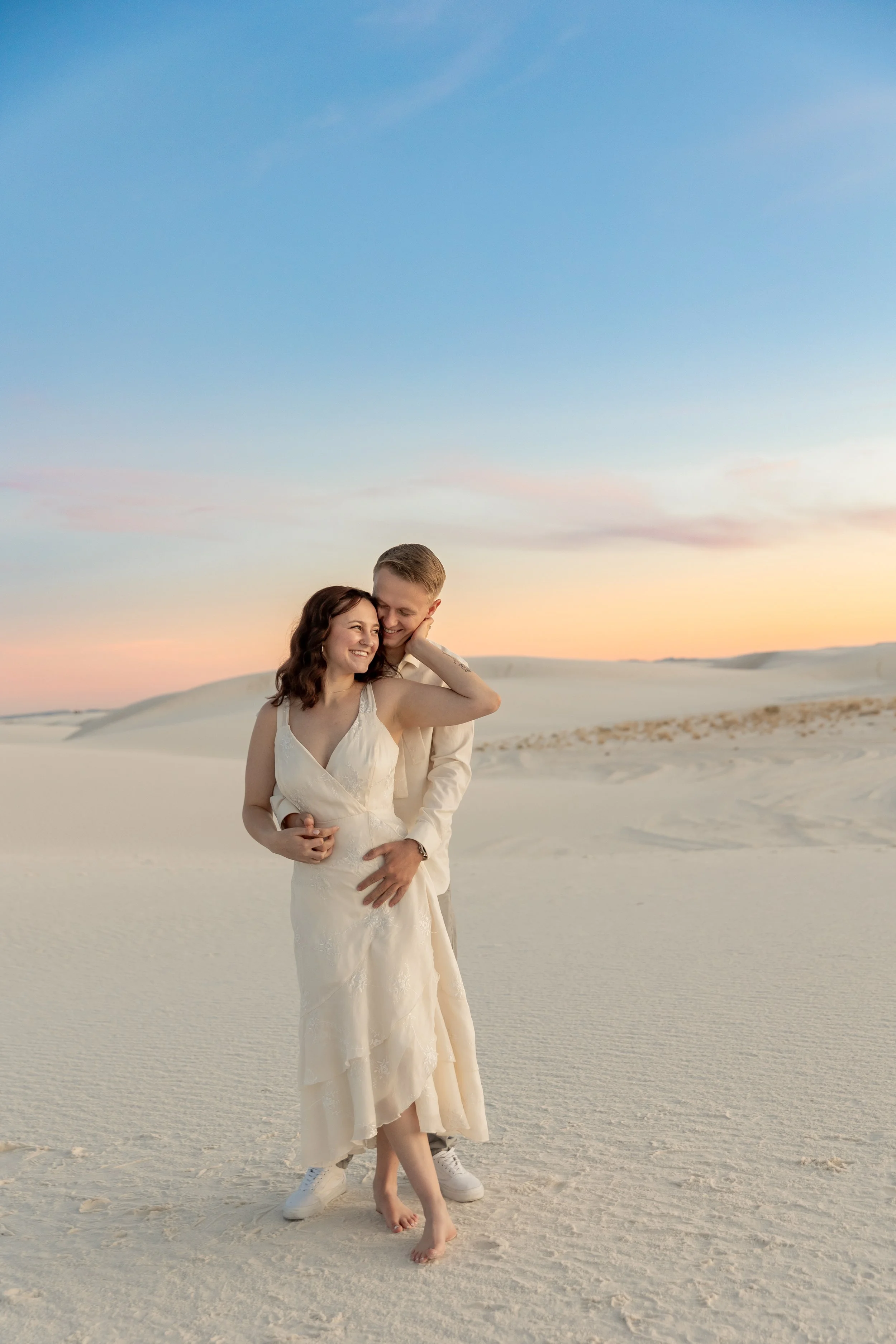 A couple in engagement attire standing barefoot on a sandy desert at sunset, embracing and smiling at White Sands National Park New Mexico