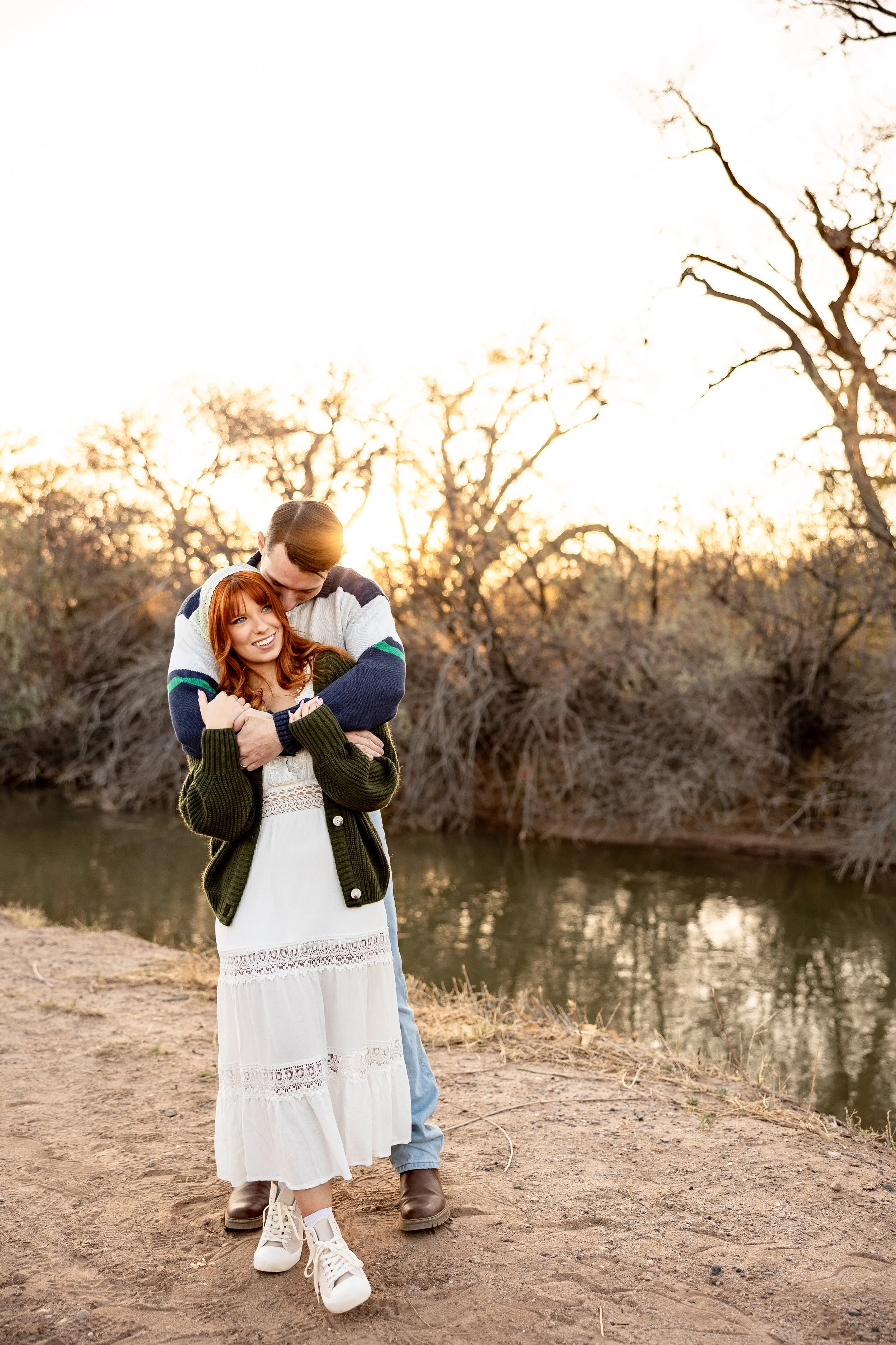 A couple embracing near a river at sunset, with trees in the background and a woman wearing a white dress and a green cardigan for an engagement in Albuquerque, New Mexico