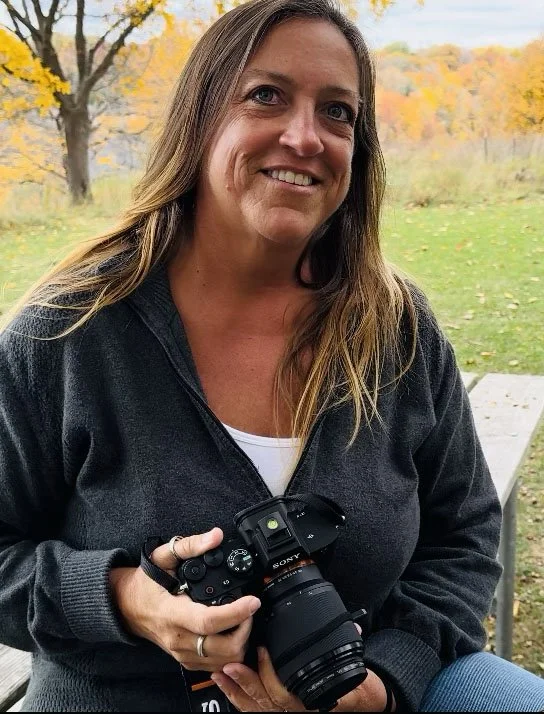 A woman smiling outdoors holding a camera, with trees and autumn leaves in the background.