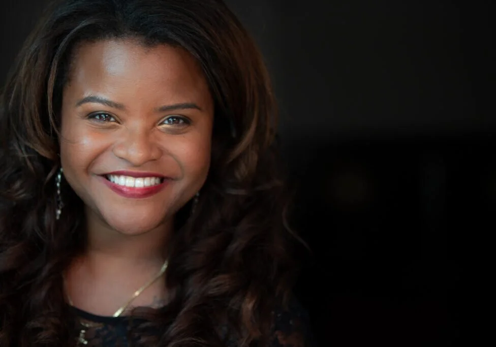 A smiling woman with long, wavy dark hair and light skin, wearing earrings and a necklace, in front of a dark background.