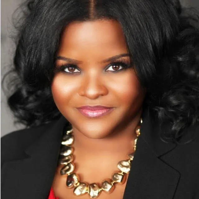 A woman with dark, curly hair smiling at the camera, wearing a black blazer and a chunky gold necklace.
