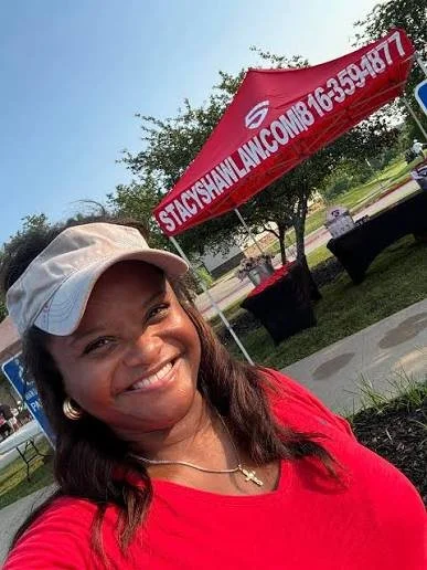 Smiling woman wearing a white cap and red shirt standing outdoors near a red banner and trees.