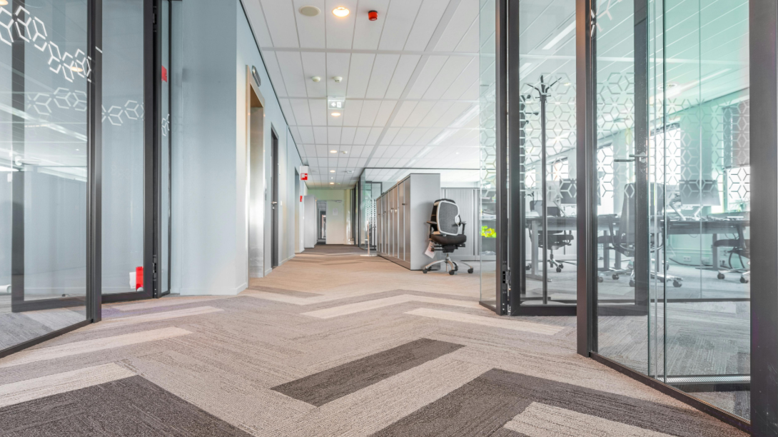 Empty modern office corridor with glass-walled conference rooms on either side, office chairs, and carpeting.