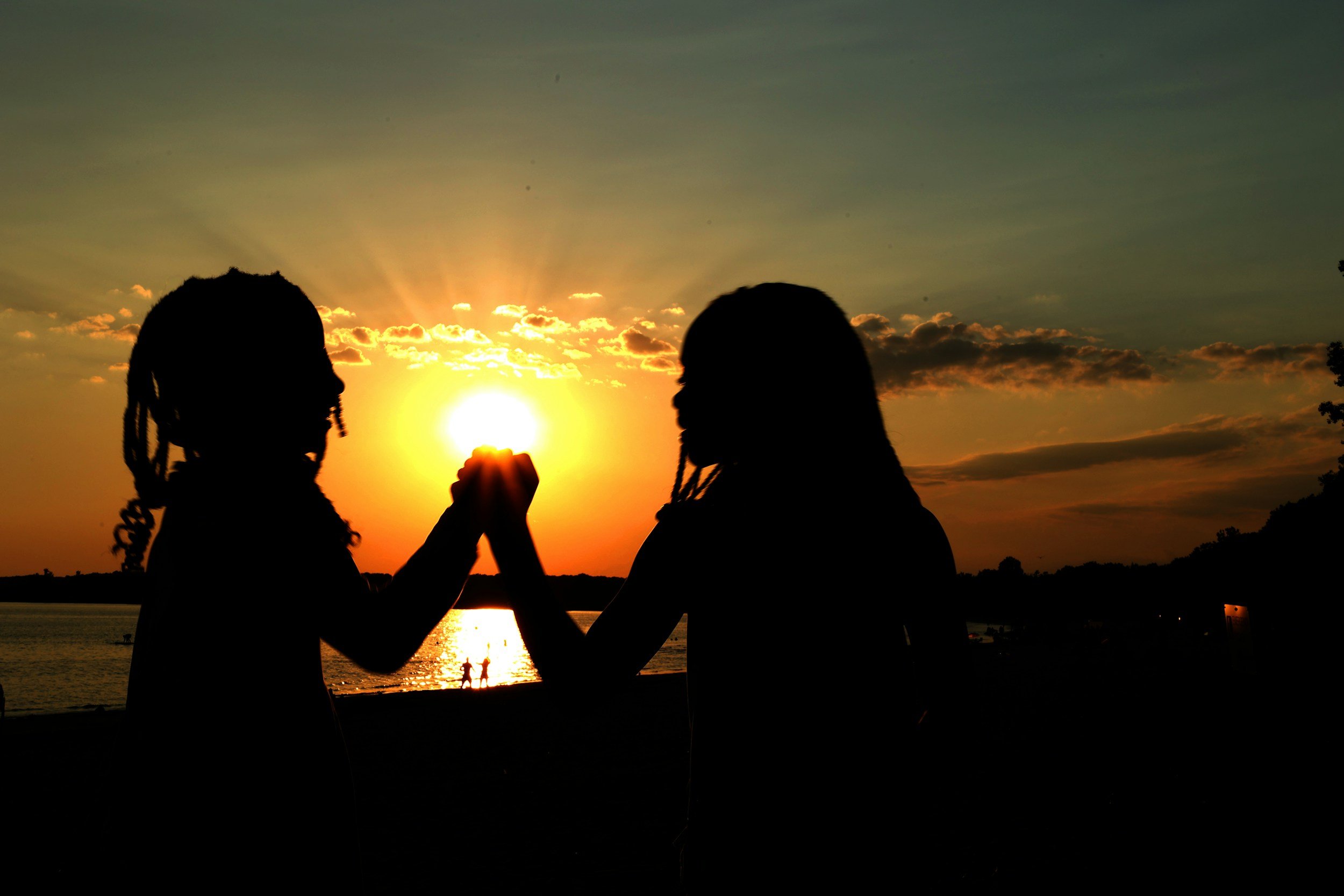 Two silhouetted women facing each other and holding hands at sunset by the water, with the sun low on the horizon and a few other people in the background near the shoreline.