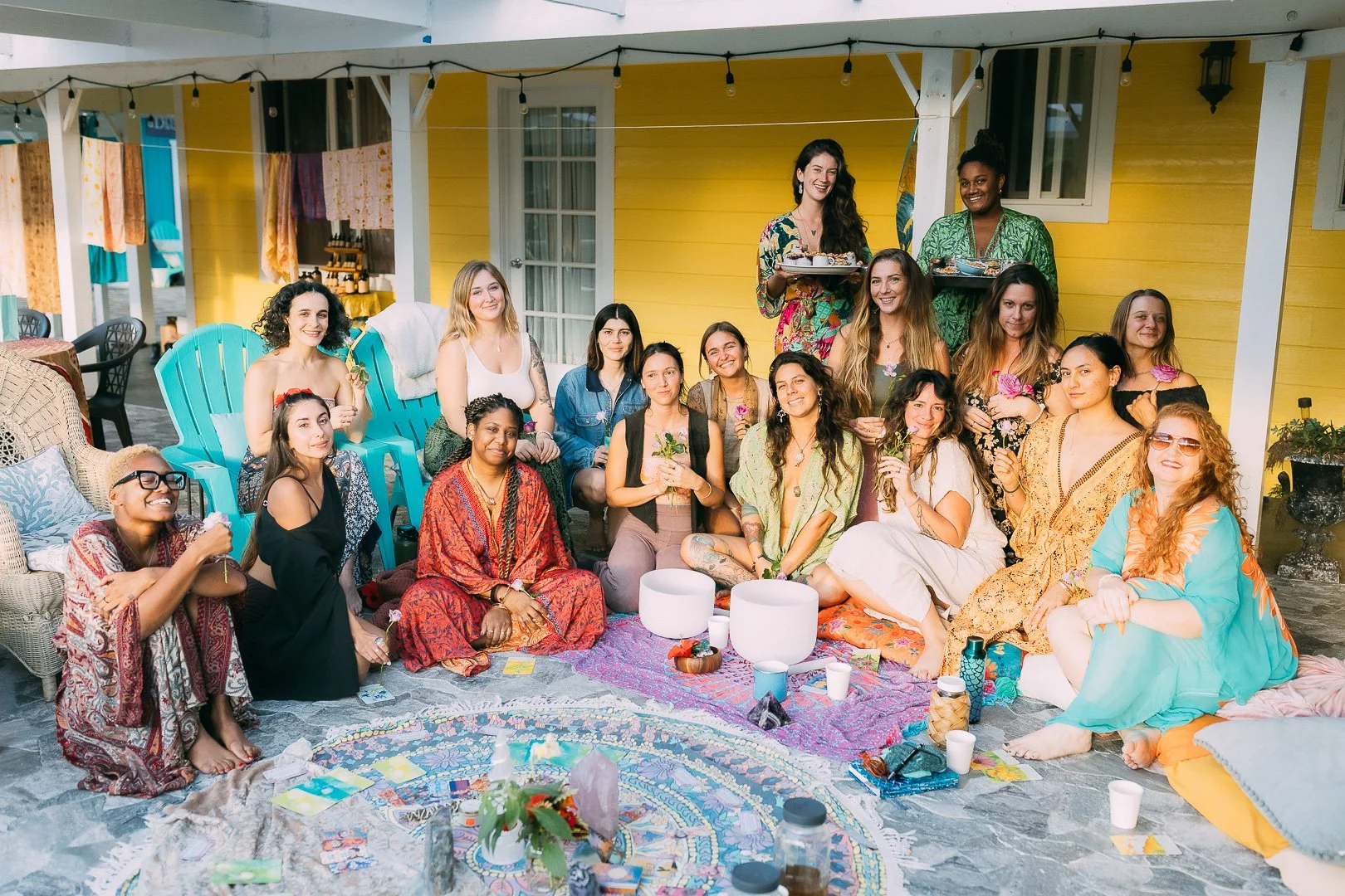 A group of diverse women gathered outdoors on a porch for a celebration, sitting on colorful rugs and pillows, with some standing behind, smiling and holding flowers and beverages.