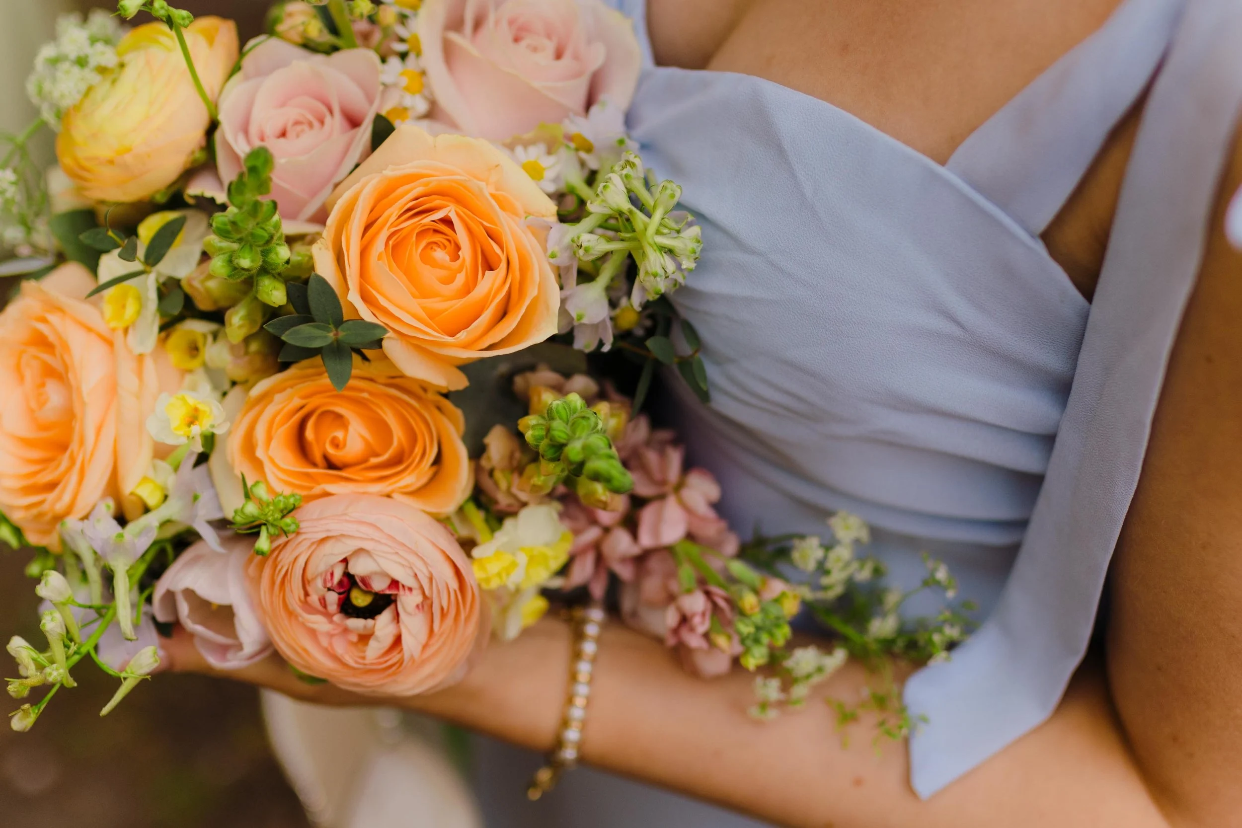 A bridesmaid holding a large bouquet of peach, pink, and white flowers, including roses, ranunculus, and small blossoms, near their chest, wearing a light blue dress