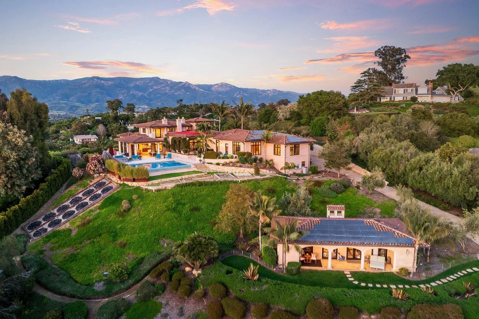 Aerial view of a luxury hilltop estate with a villa and pool surrounded by lush greenery and mountains in the background, under a sunset sky.