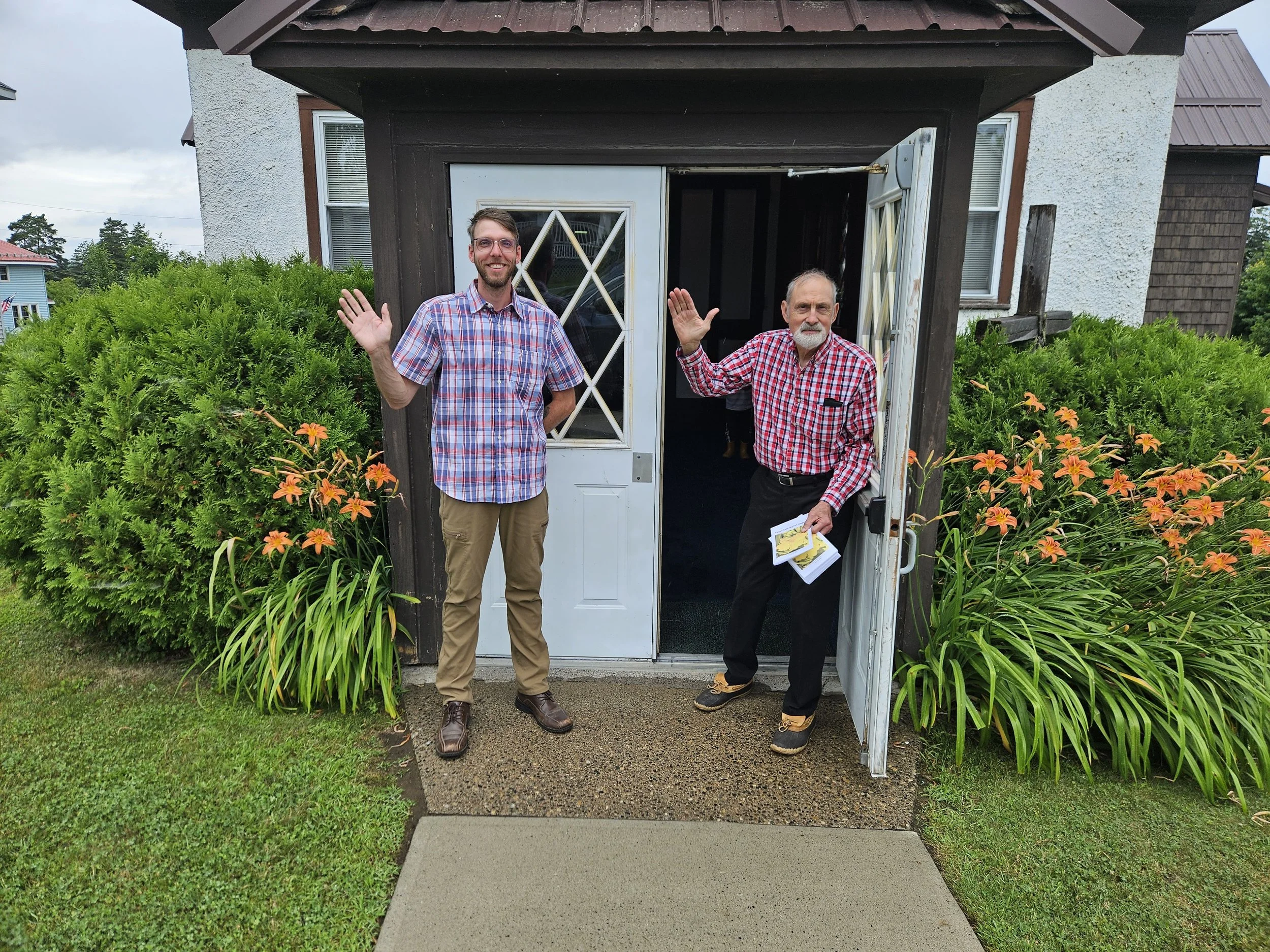 Two men standing outside a house, waving. The younger man is on the left, wearing a plaid shirt and khakis. The older man is on the right, wearing a red plaid shirt and black pants, holding papers in his left hand and waving with his right hand. They are in front of a white door surrounded by green bushes with orange flowers.