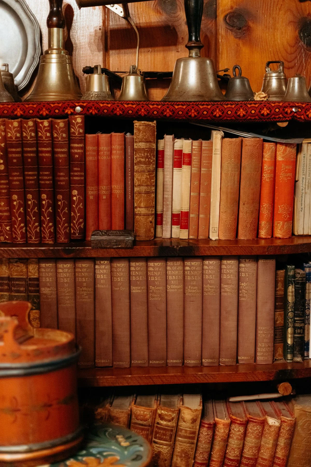 A wooden bookshelf filled with various antique books, some with red and beige covers, and decorative items including bells and a metal tray on top.