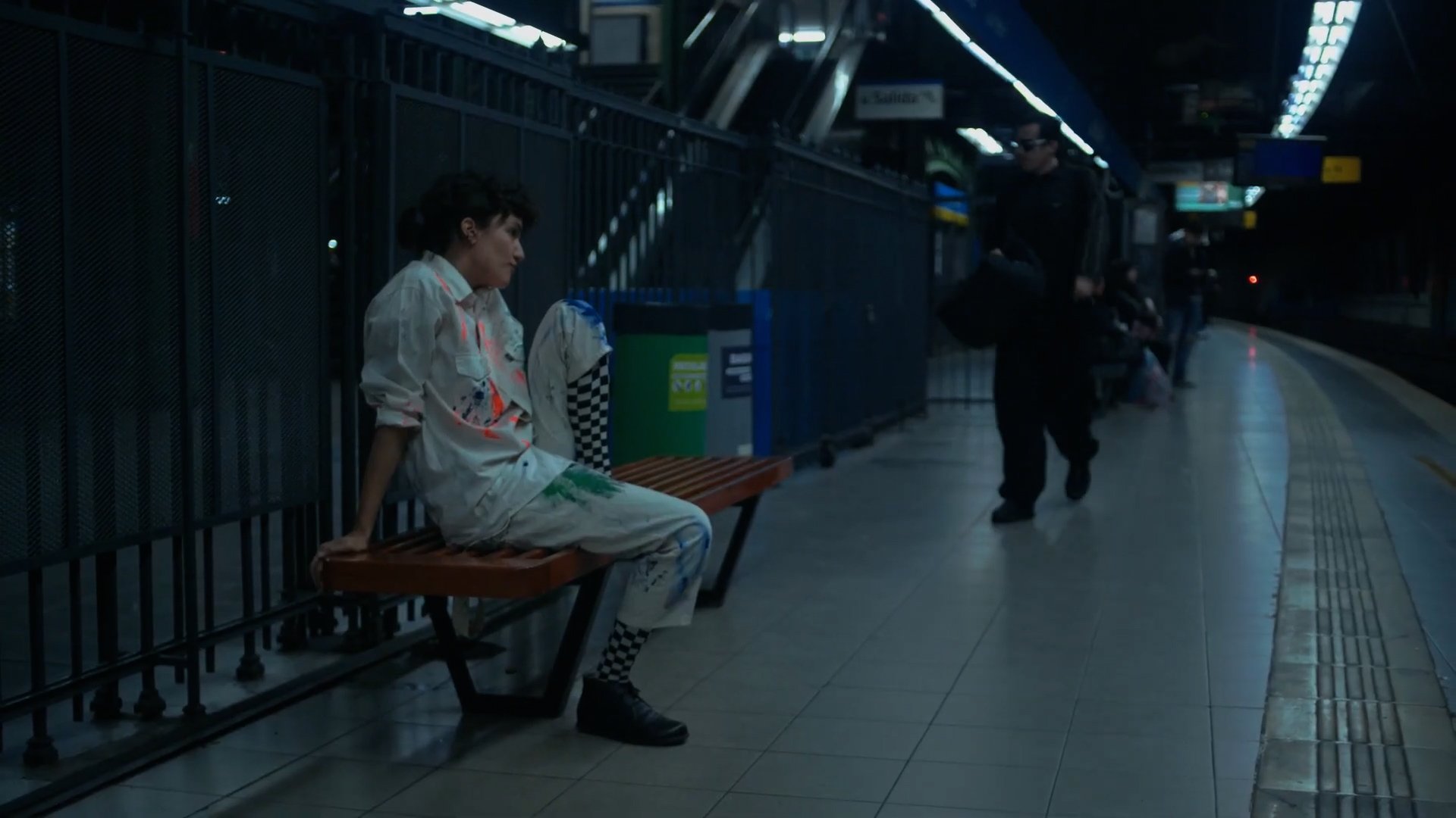 Planta x NAP SPOT. Persona sentada en un banco de una estación de metro desierta, vestida con ropa clara y calcetines a cuadros. Otra persona camina por el andén.