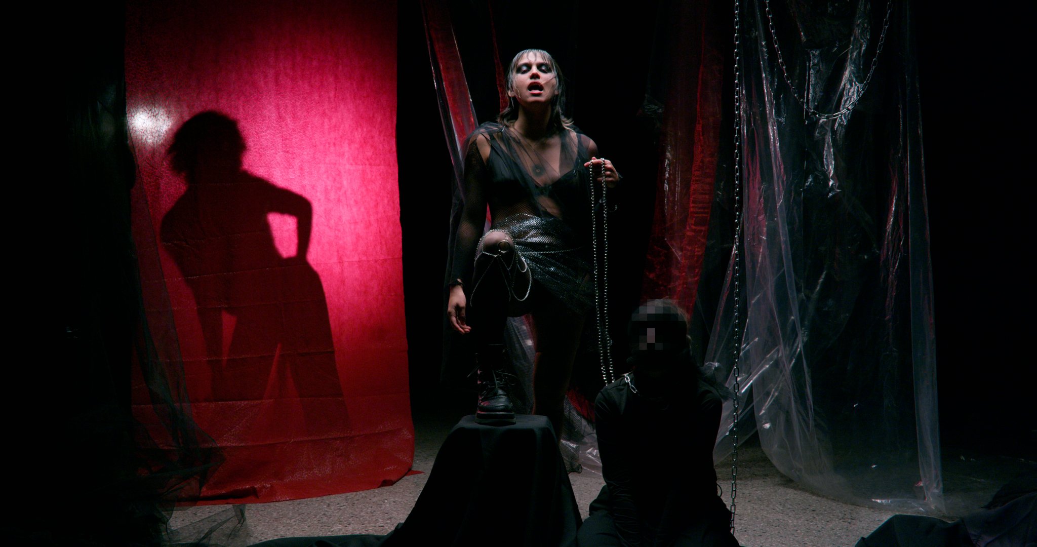 Mainé. Persona en escenario oscuro con cortinas rojas, sosteniendo una cadena. Sombra proyectada detrás. Ambiente teatral y dramático.