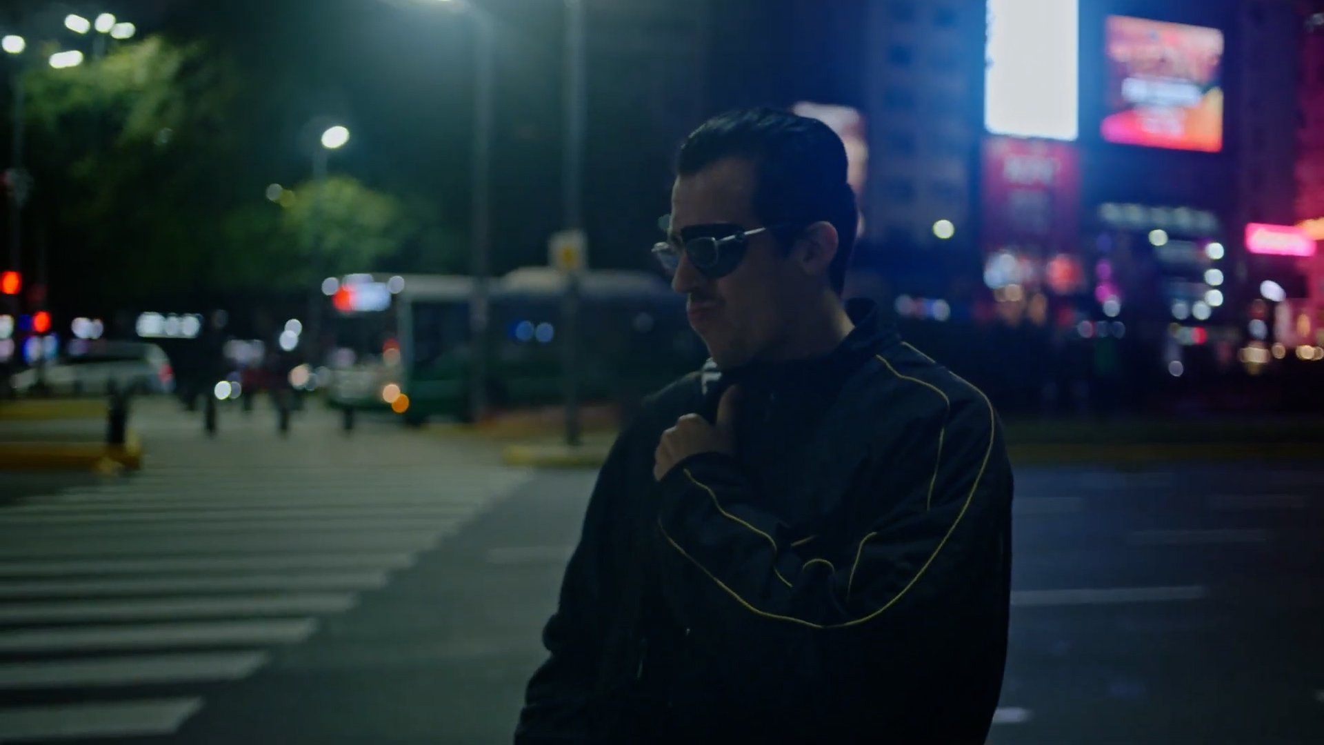 Planta x NAP SPOT. Persona con gafas de sol caminando por una ciudad por la noche, con fondo urbano que incluye luces brillantes y un paso de cebra.