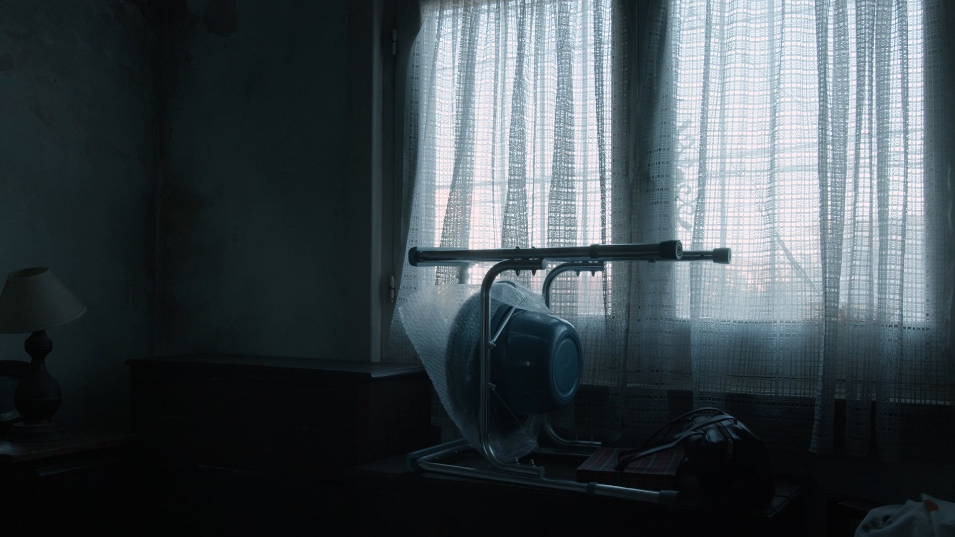 La Higuera. Habitación oscura con una ventana cubierta por cortinas de encaje, una lámpara pequeña, y objetos envueltos en plástico sobre un mueble.