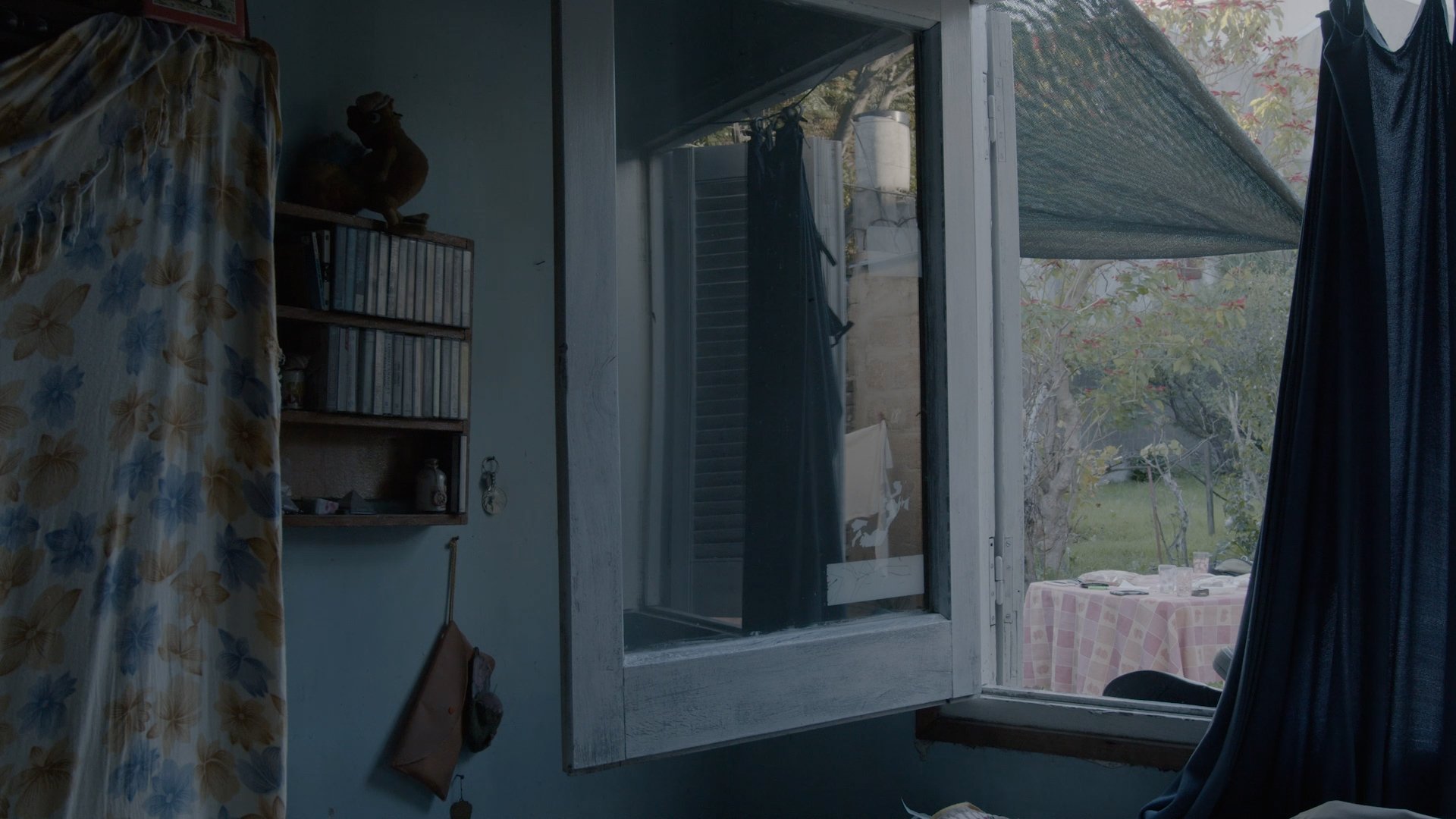 La Higuera. Interior de una habitación con ventana abierta, cortinas oscuras, una estantería con libros, y vista al exterior con un jardín y mesa con mantel a cuadros.