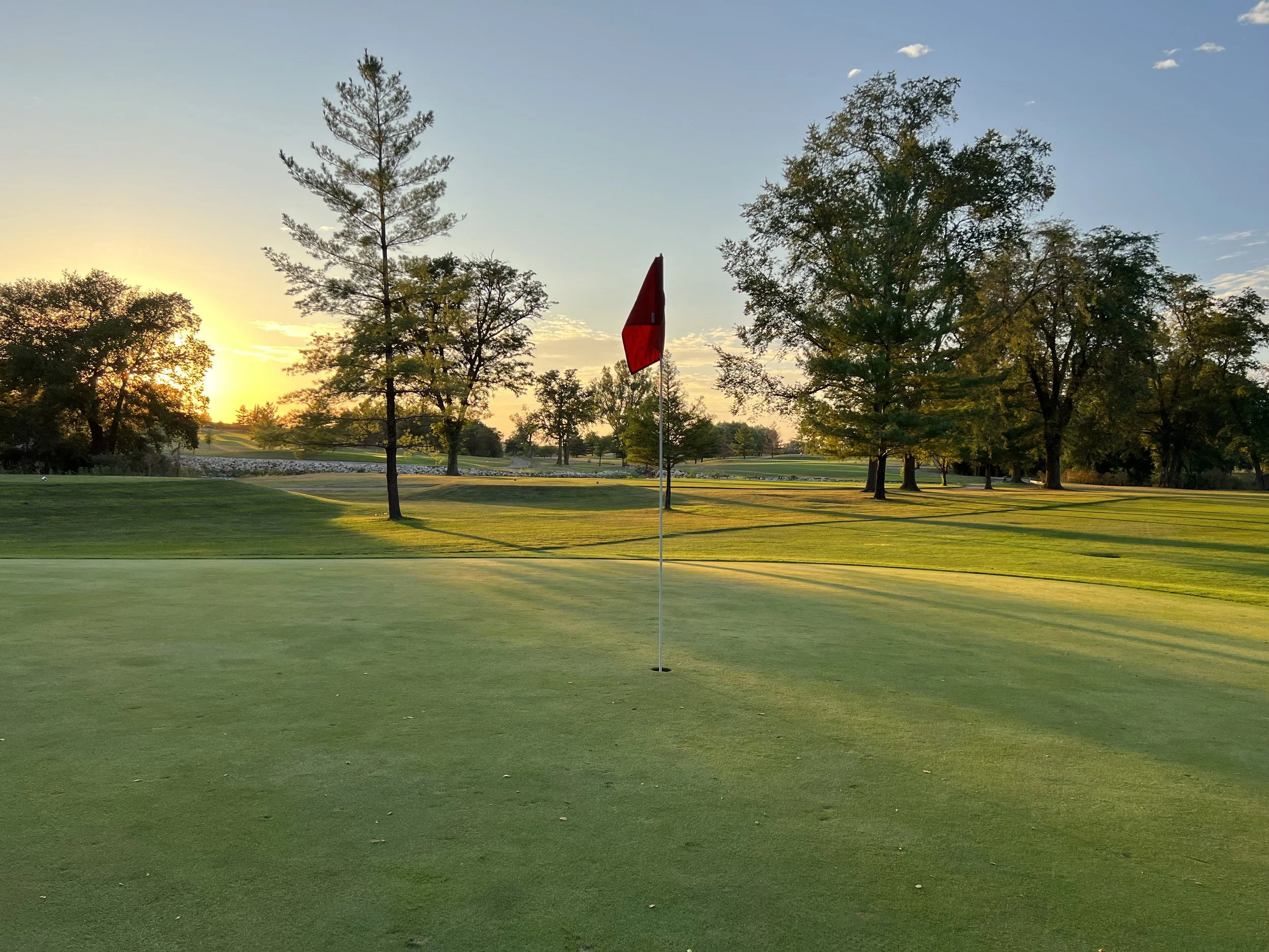 Golf course green with red flag, trees in background, sunset view