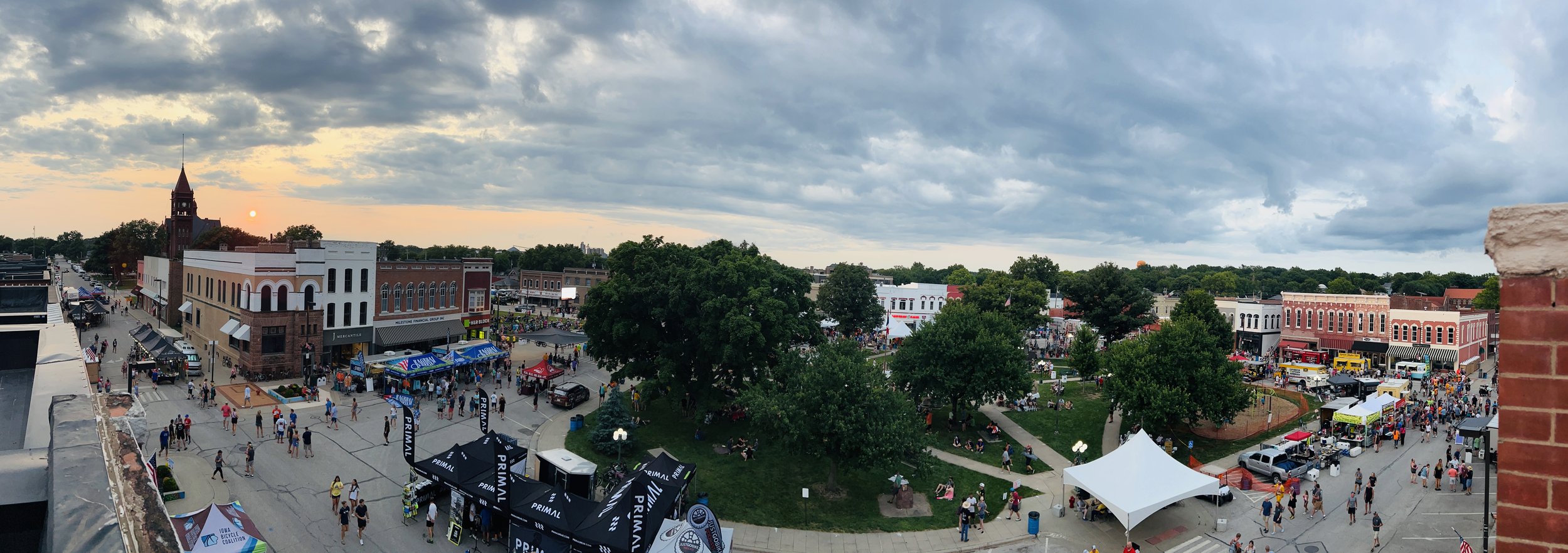 Outdoor festival in a small town square at sunset, featuring food trucks, vendor tents, trees, and a clock tower, with numerous people attending the event.