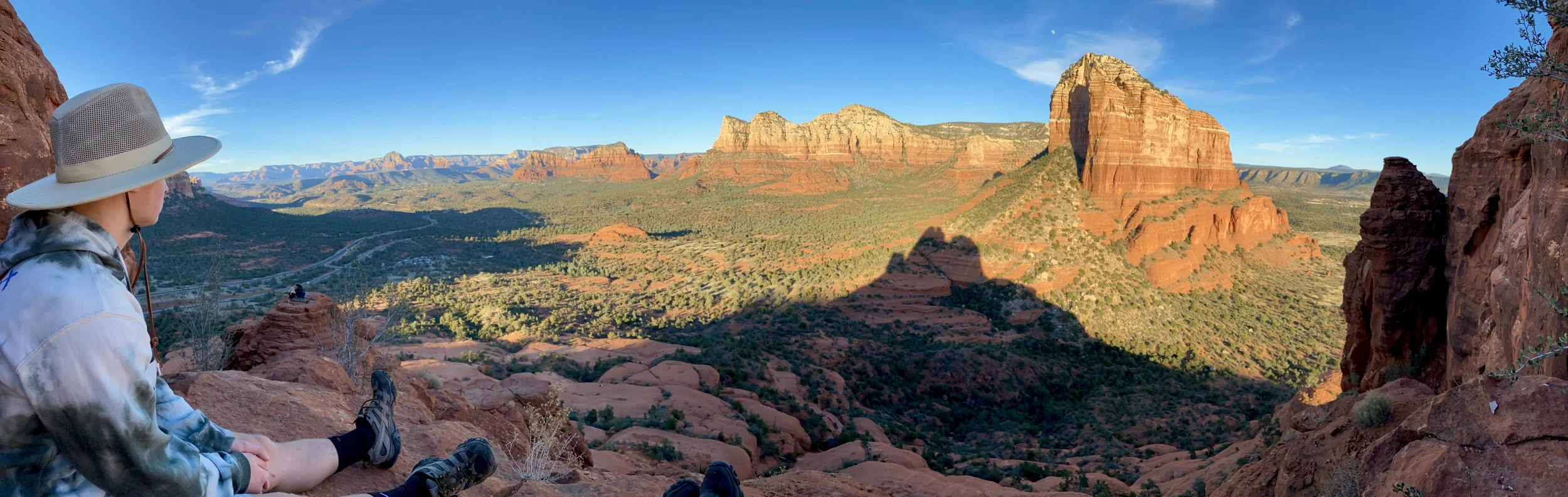 Person sitting on a rocky ledge overlooking a panoramic view of red rock formations and a desert valley under a clear blue sky.