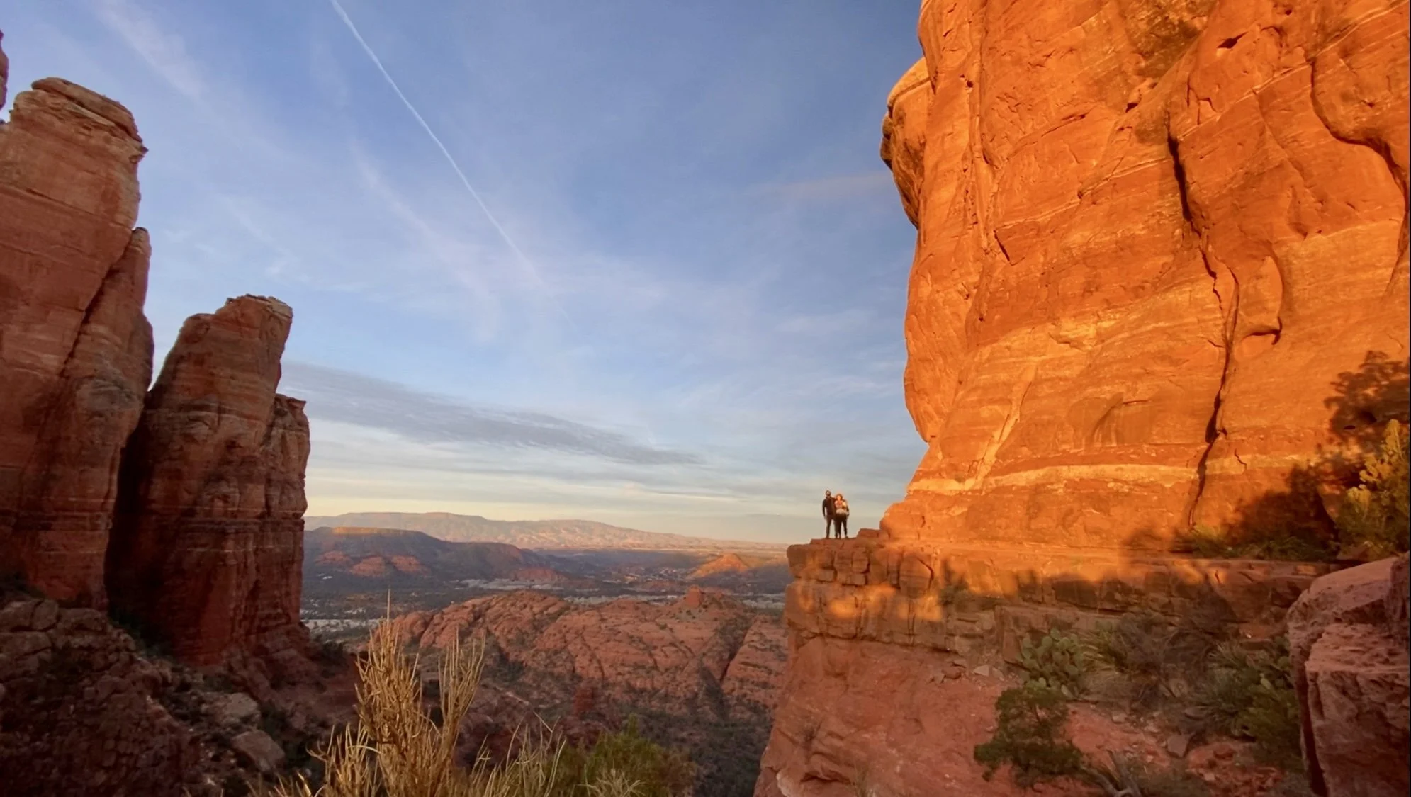 Two people standing on a red rock cliff with expansive views of Sedona's rugged landscape and distant mountains, under a clear blue sky.