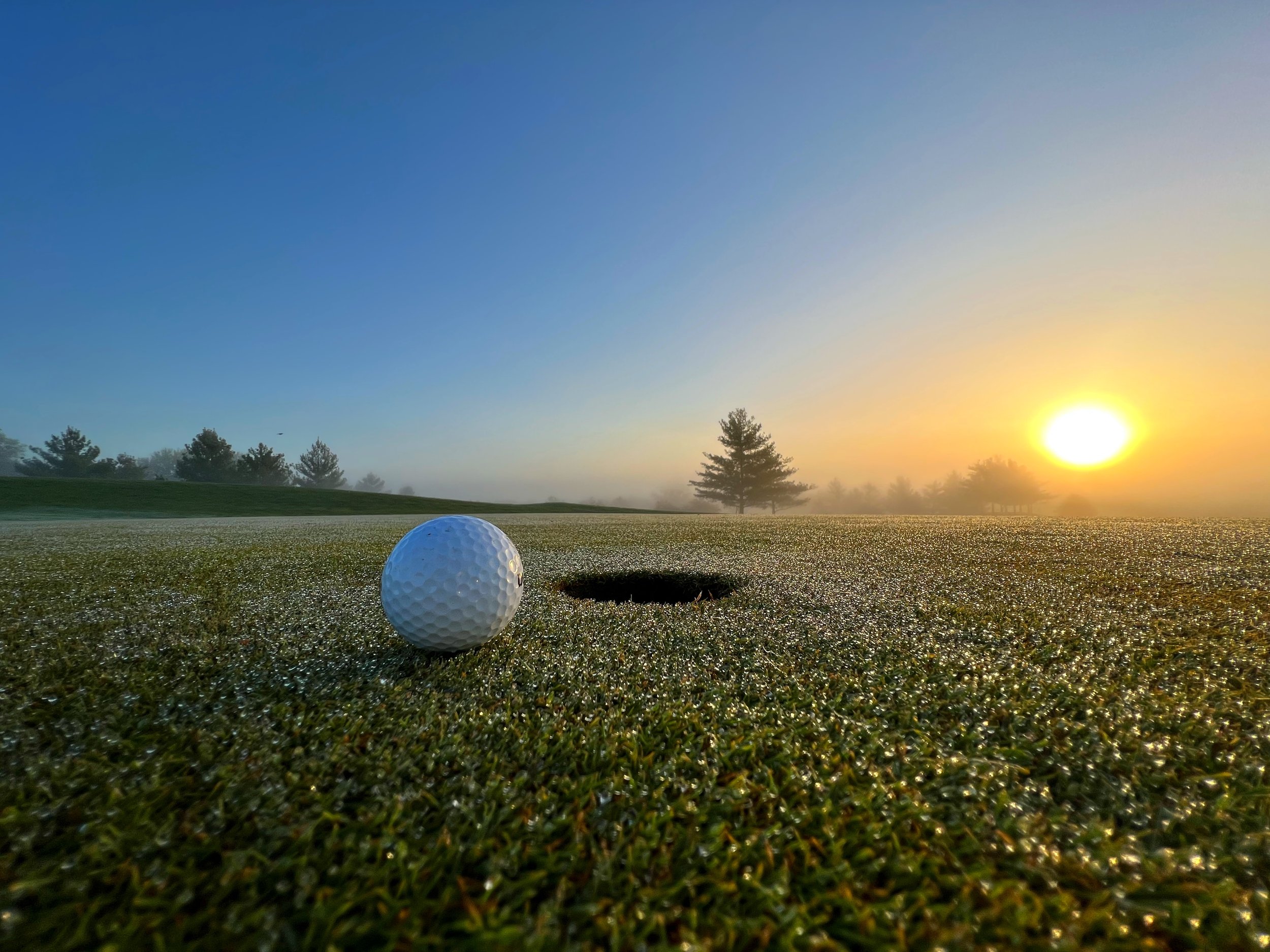 Golf ball on a dewy green near the hole with sunrise in the background.