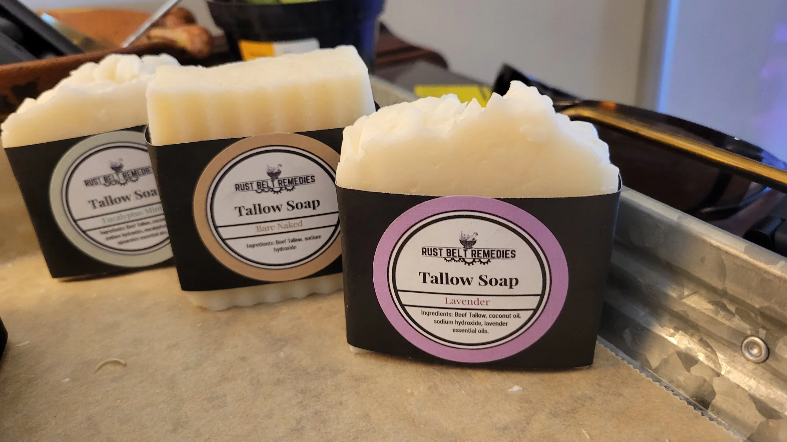 Three bars of handmade soap labeled as Tallow Soap from Rust Belt Remedies, with scents of Bare Naked and Lavender, displayed on a countertop.
