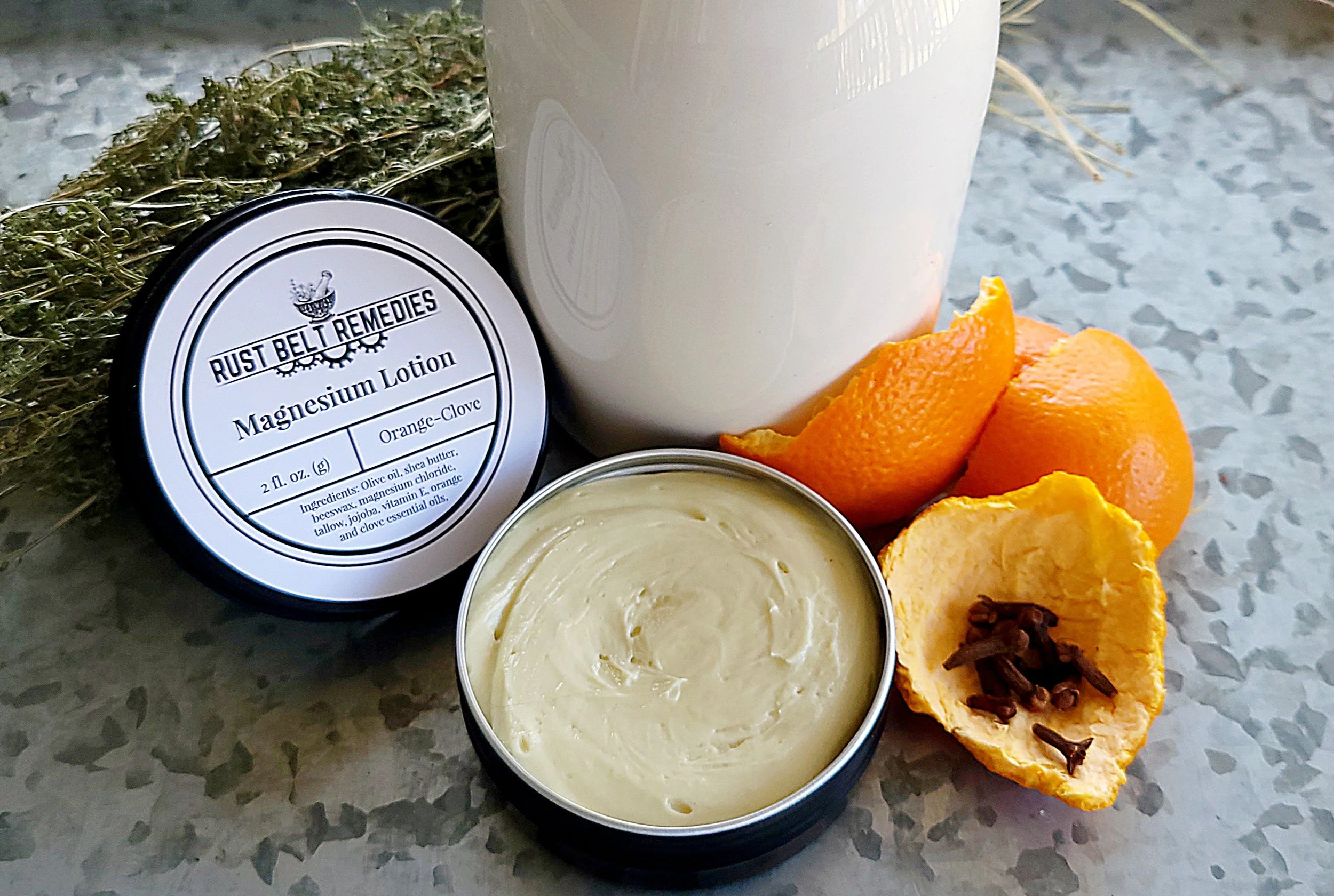 A white container of Rust Belt Remedies Magnesium Lotion, a small open tin of cream, two oranges, a halved orange with cloves inside, and a bundle of dried herbs on a speckled gray surface.