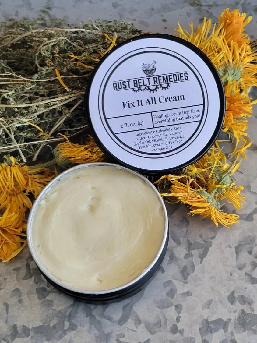 A tin container of Fix It All Cream from Rust Belt Remedies with dried yellow flowers and green herbs underneath, and the lid leaning against it.