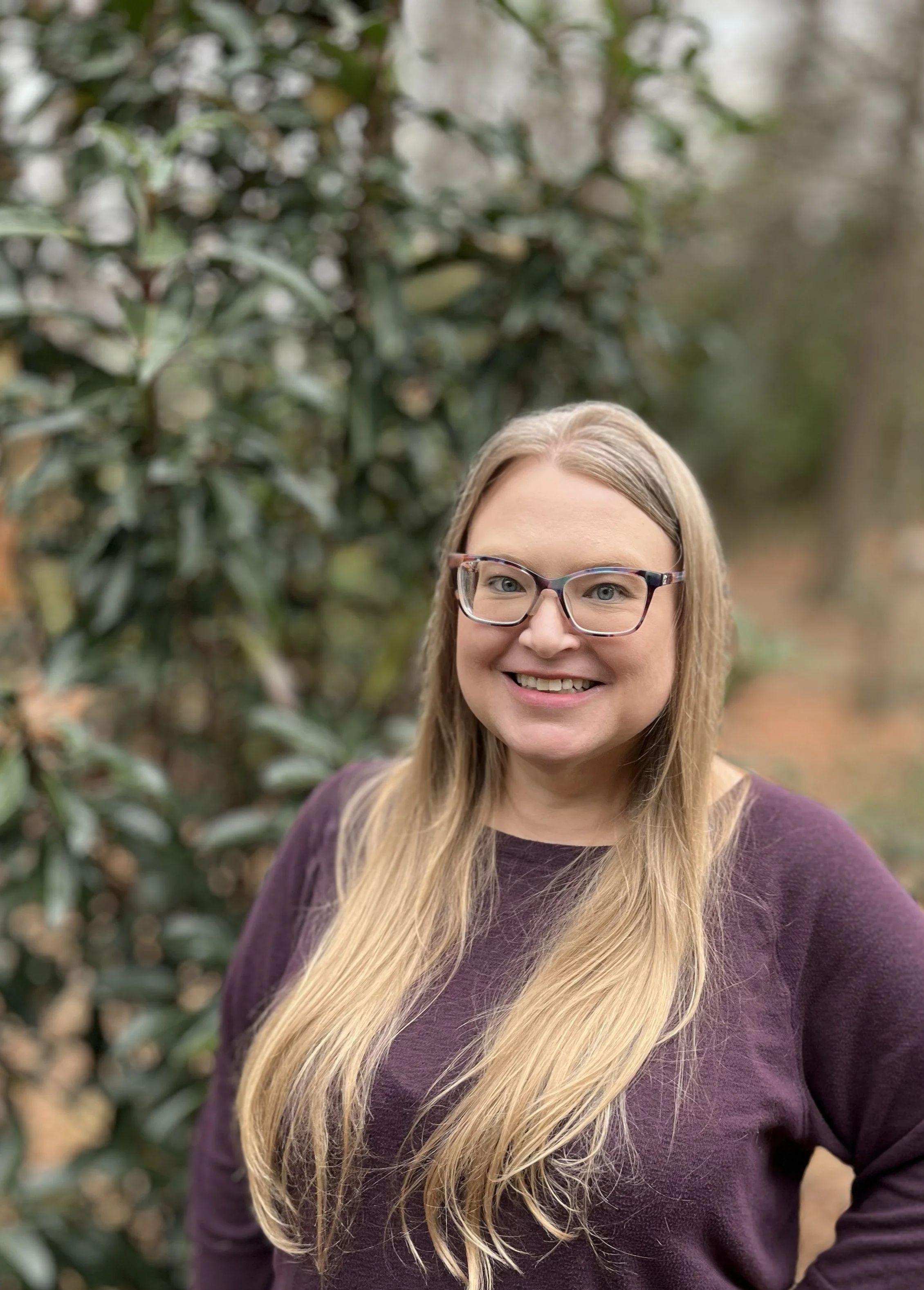 Lifespan Religious Education Coordinator Jennifer smiles and stands in the foreground of a wooded scene