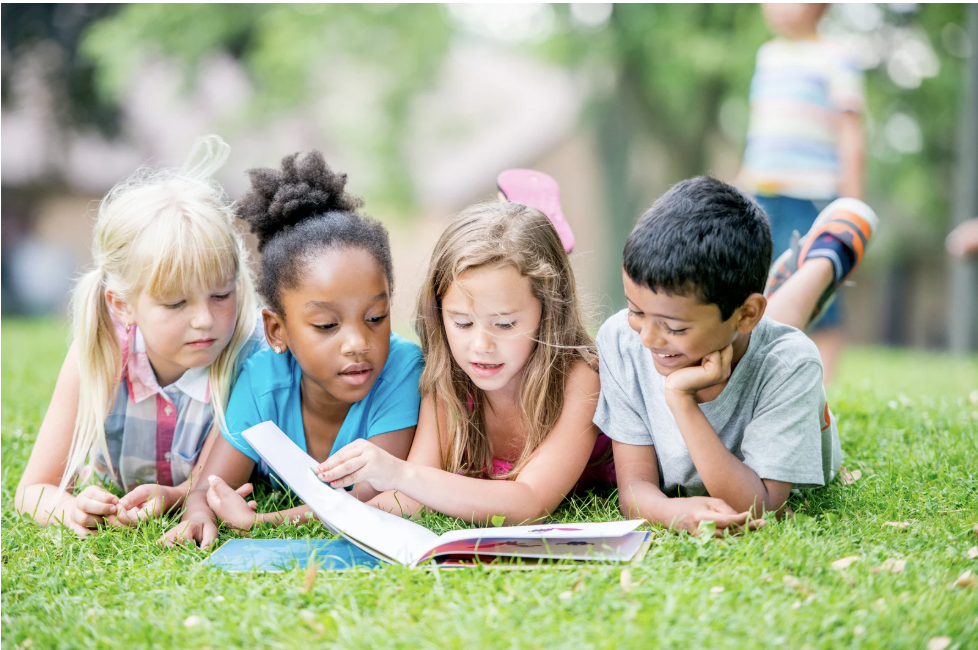 Four children lying on grass reading a book together.