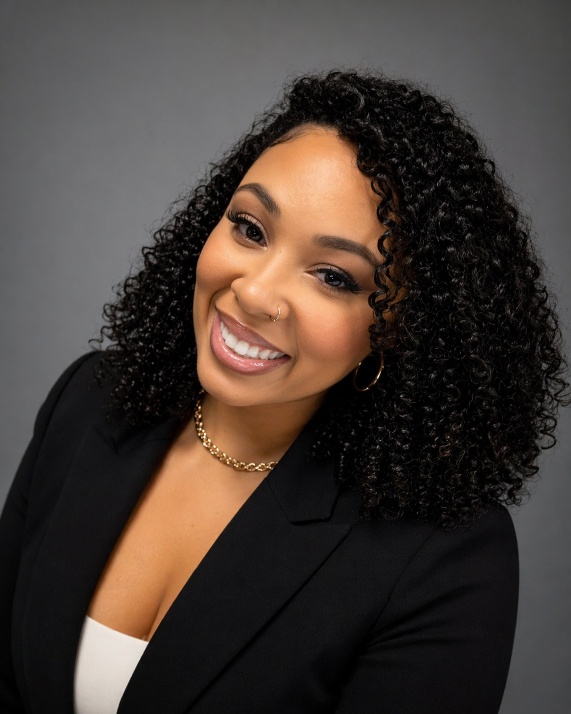 Portrait of a smiling woman with curly black hair, gold hoop earrings, a nose ring, and wearing a black blazer and gold necklace against a gray background.