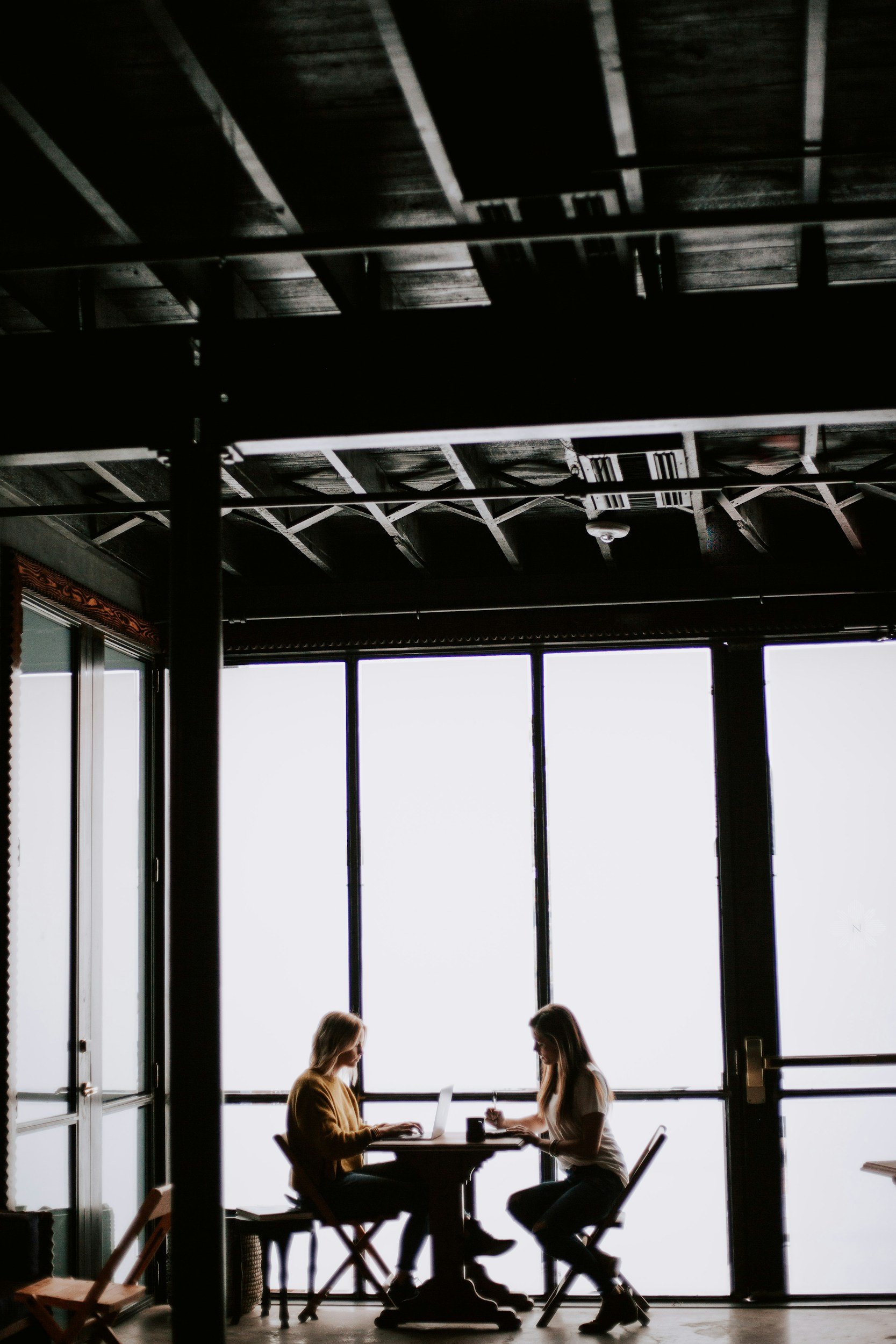 Two women sitting at a table in front of large windows, working on laptops and taking notes, inside a modern building with exposed ceiling beams.
