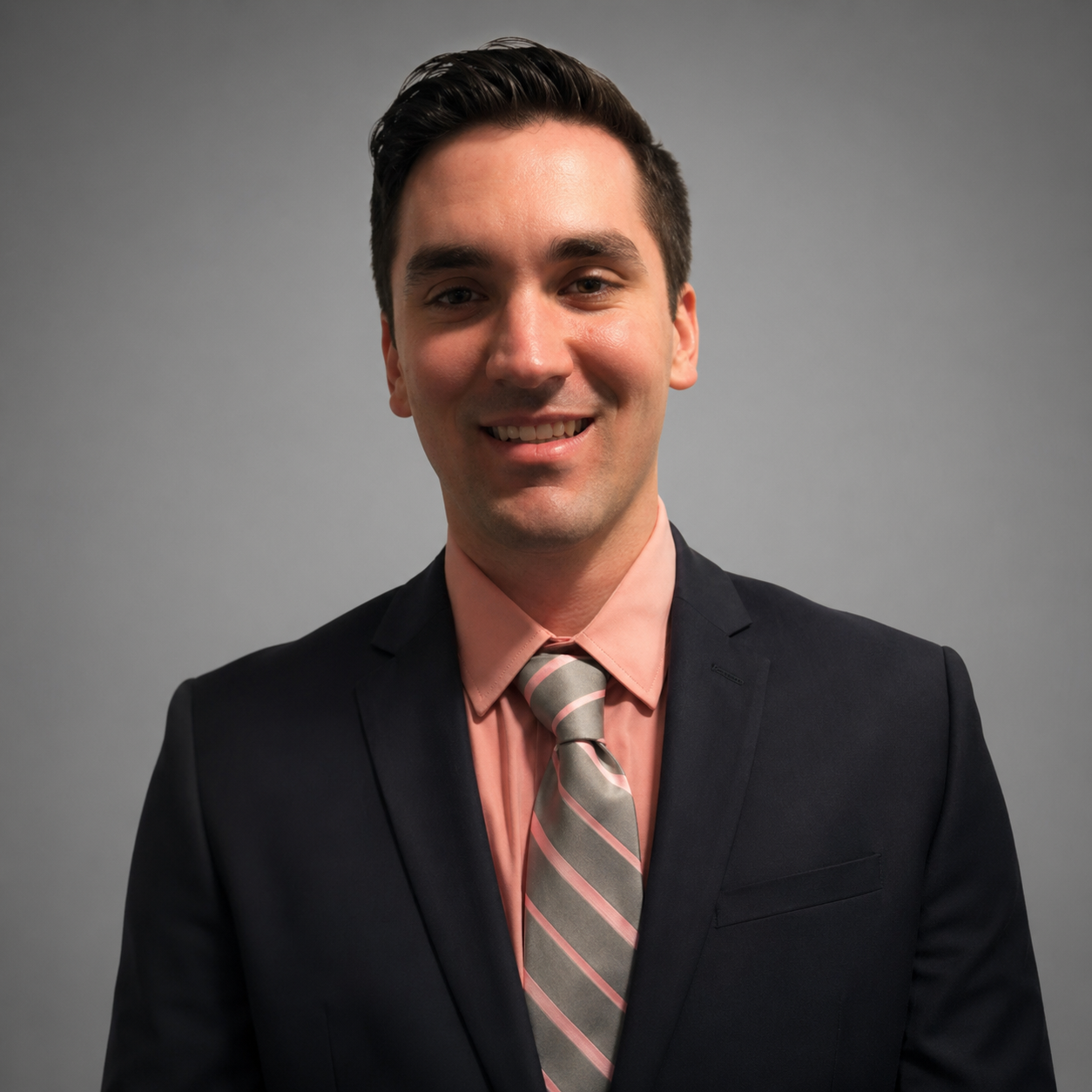Portrait of a young man with dark hair, smiling, wearing a dark suit, pink dress shirt, and striped pink and gray tie, standing against a gray background.