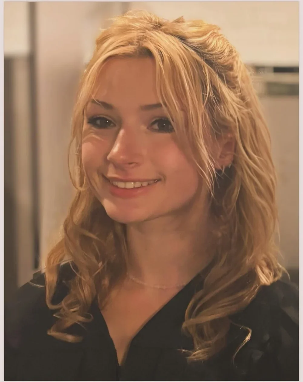 Close-up of a smiling woman with blonde curly hair, wearing a black top, in an indoor setting.