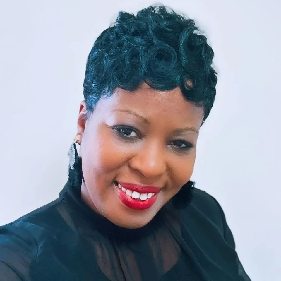 A woman with short, curly black hair, wearing a black top, smiling with red lipstick, against a plain white background.
