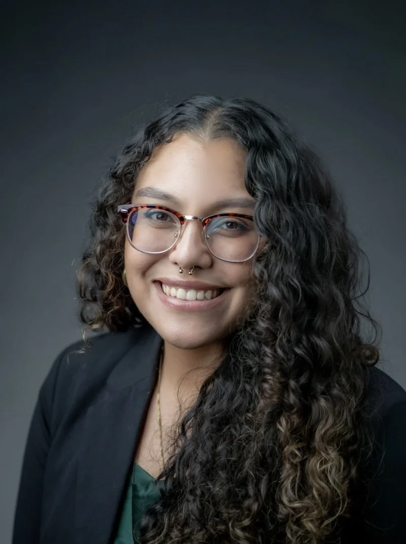 A woman with long curly hair wearing glasses, a septum piercing, and a dark blazer smiling against a dark background.