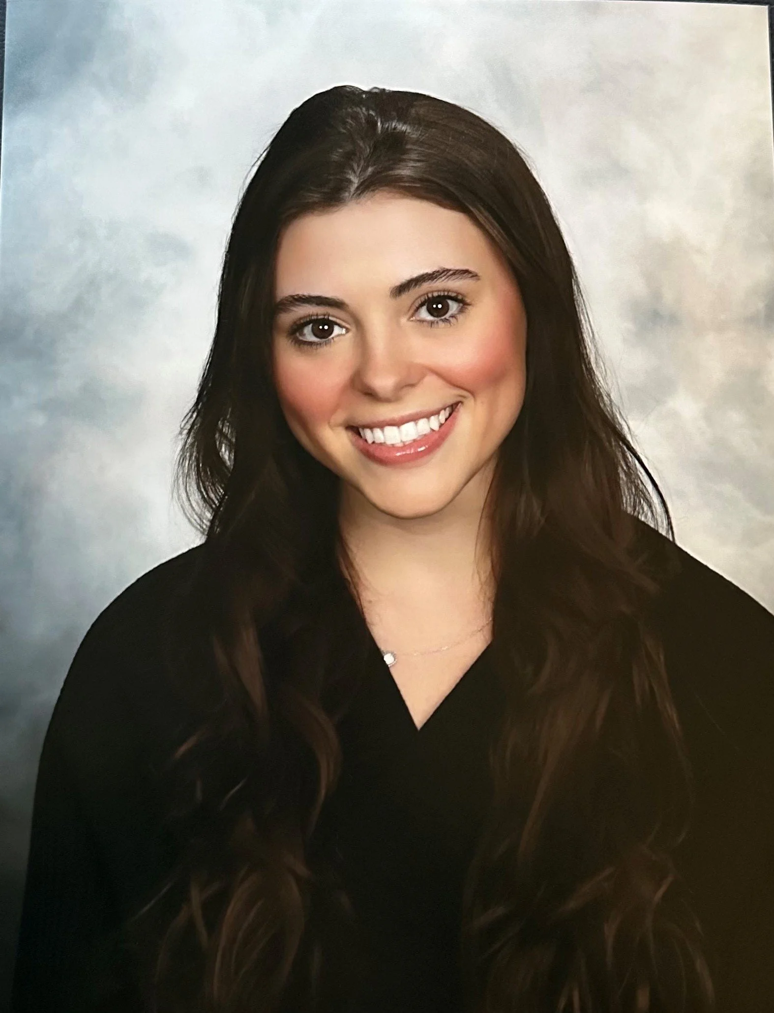 A young woman with long wavy brown hair and a bright smile, dressed in a black top, posing for a portrait against a cloudy background.