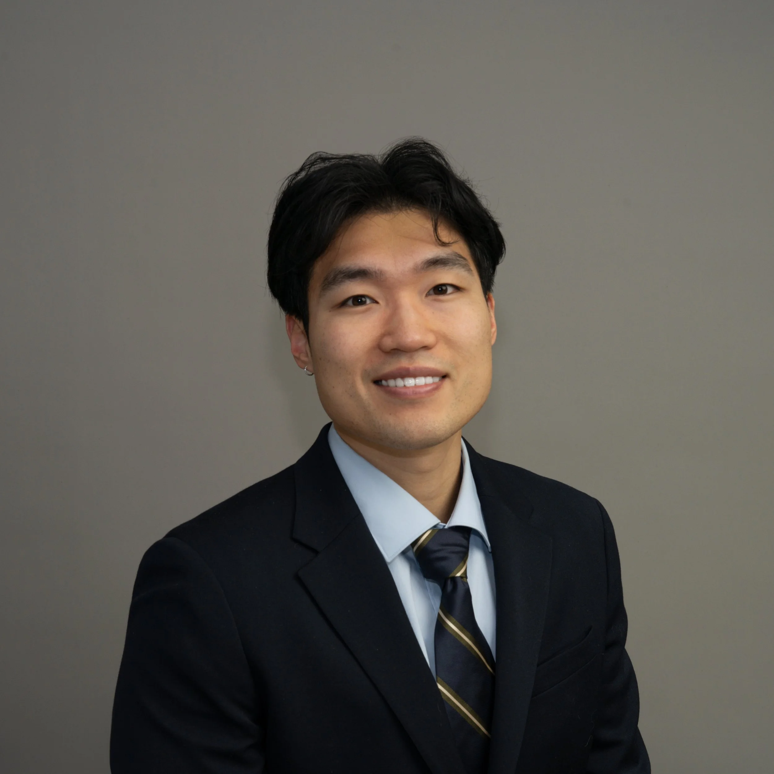 A young Asian man in a black suit, white shirt, and striped tie smiling against a plain grey background.
