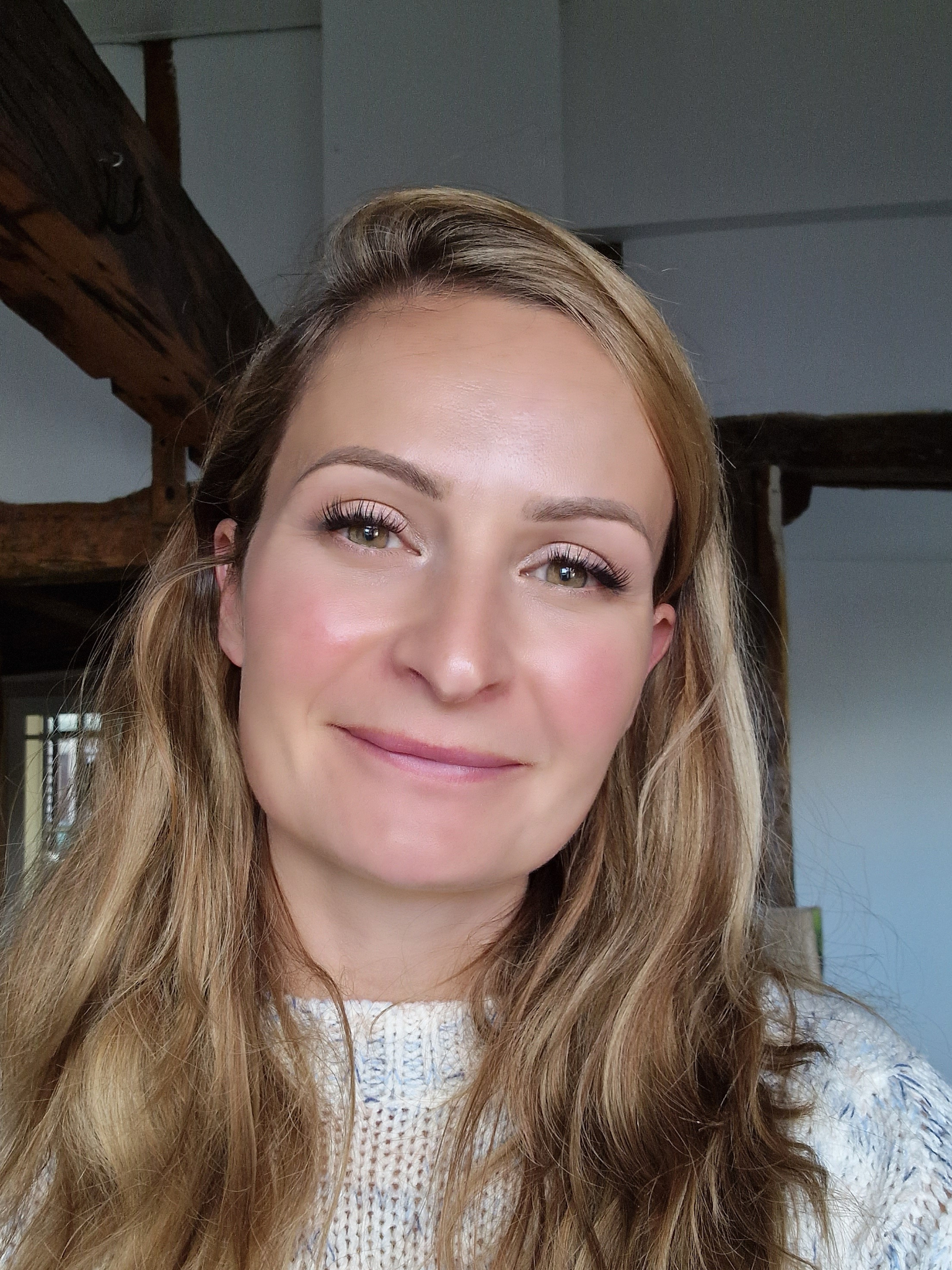 Close-up portrait of a smiling woman with long wavy blonde hair, wearing a light-colored knit sweater, indoors with wooden beams in the background.