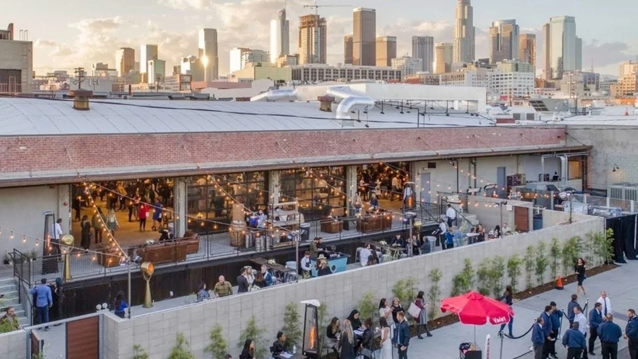 Rooftop restaurant in downtown Los Angeles with people dining, string lights, and the city skyline in the background during sunset.