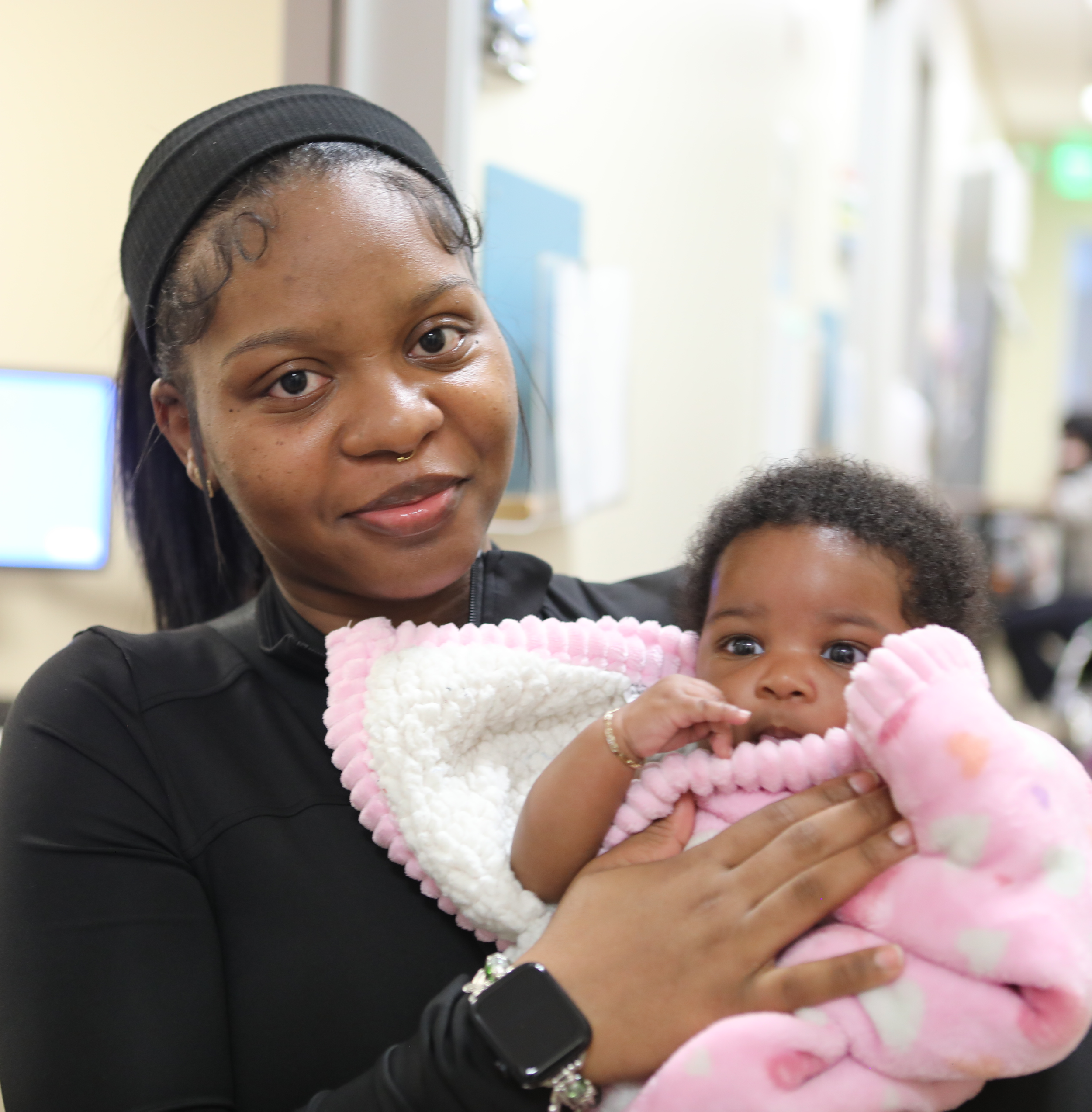 A woman with dark hair wearing a black headband and earrings holding a young child in a pink and white fluffy blanket. The woman has a nose piercing and is wearing a black top. The background shows a hospital or clinic setting.