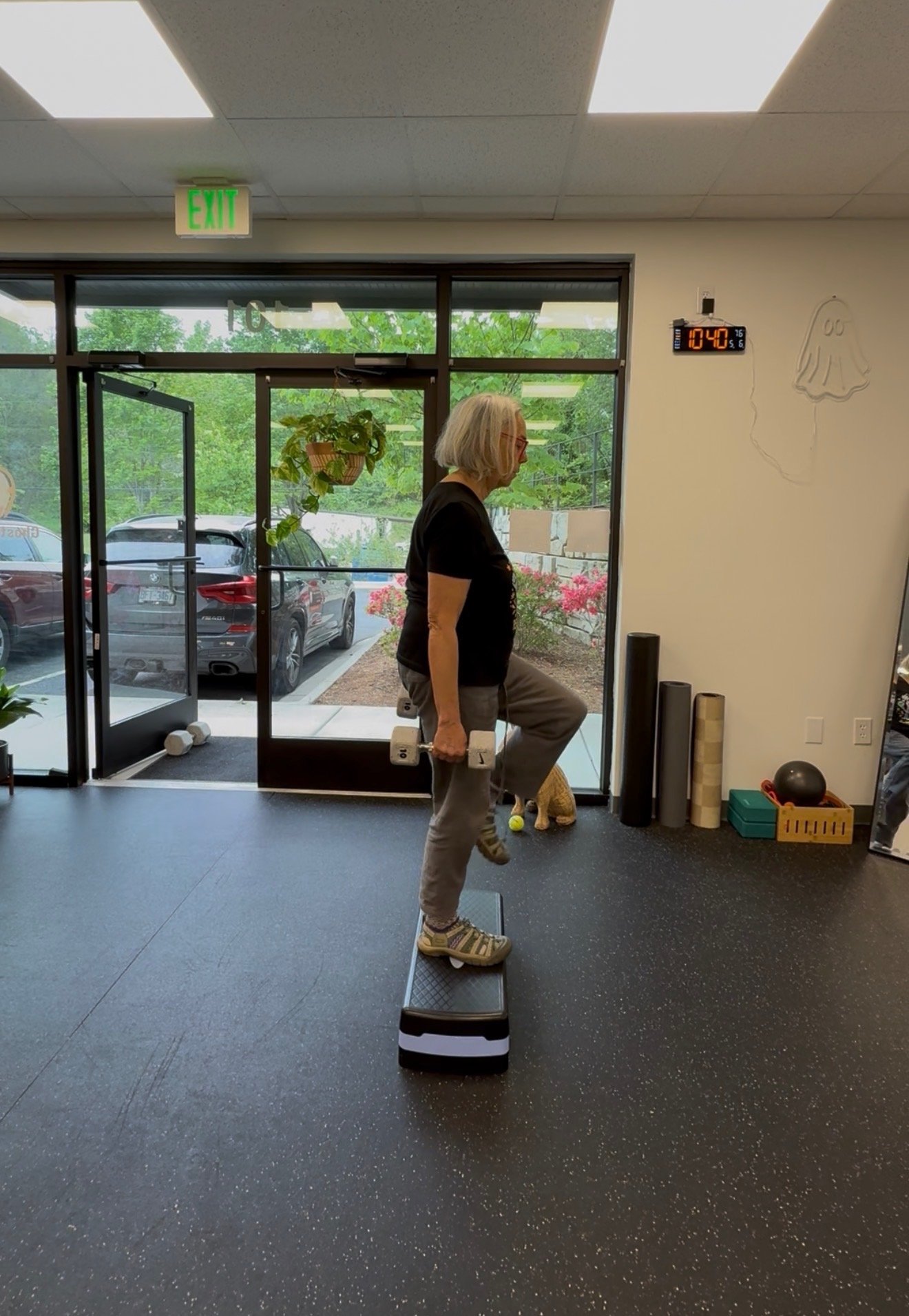 A woman doing balance exercises on a step platform during a fitness session inside a gym. She is holding dumbbells in her hands and standing on one leg while wearing workout clothes and sneakers. The gym has exercise equipment, a large window showing parked cars outside, and a digital clock displaying 10:40.