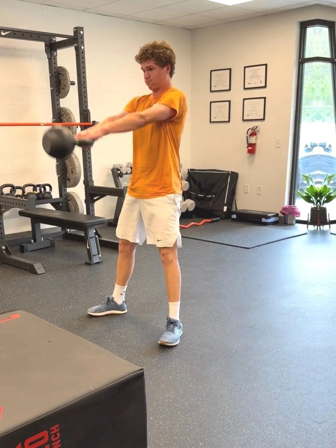A young man wearing an orange shirt, white shorts, and gray sneakers is lifting a black kettlebell with both hands in a gym. The gym has black rubber flooring, a squat rack, framed certificates on the wall, Athlete personal training. Personal trainin
