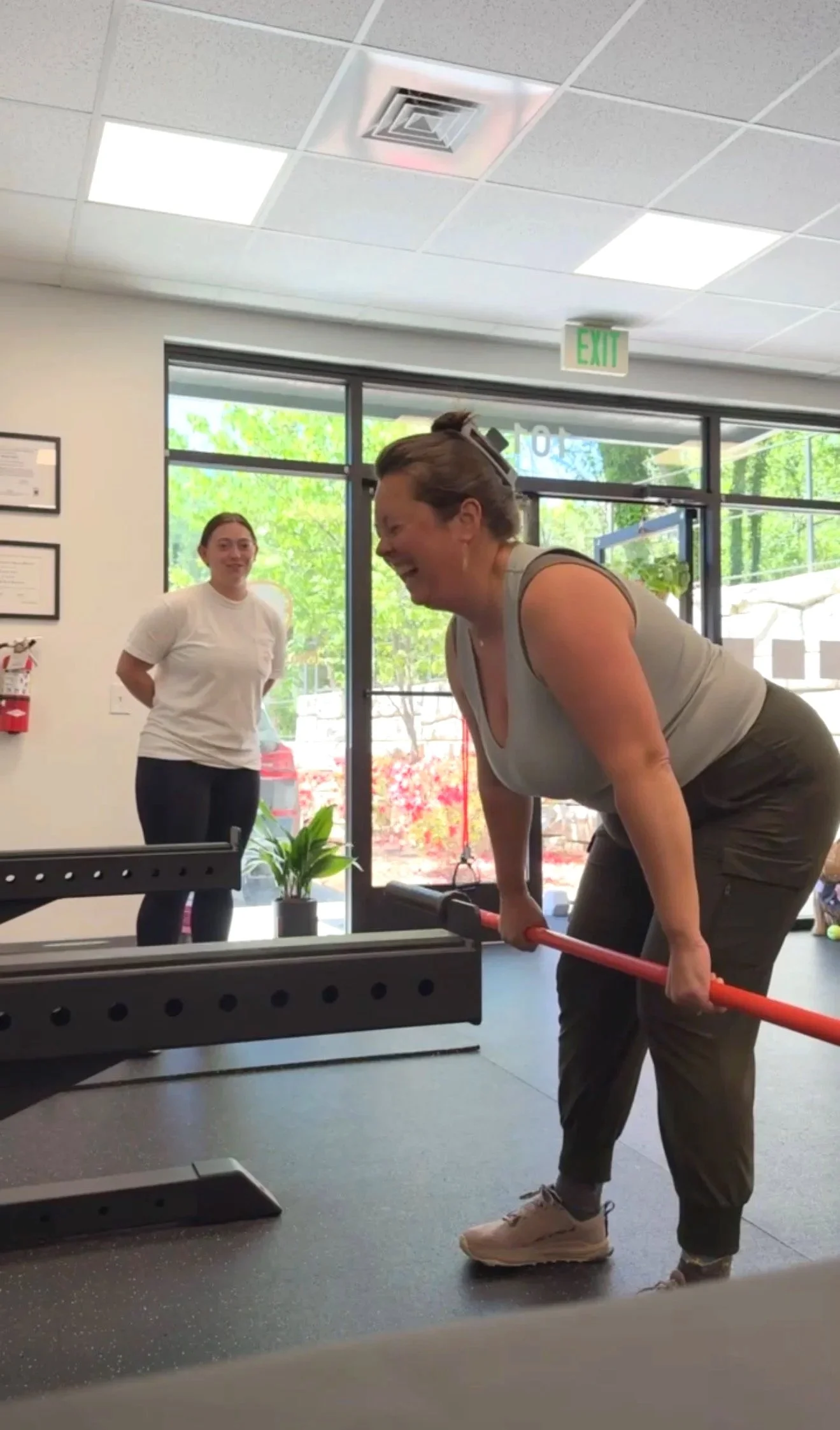 A woman with short hair in workout clothes is lifting a barbell in a gym. A woman with long hair tied back is smiling and watching in the background.Athlete personal training. Personal training Asheville NC.